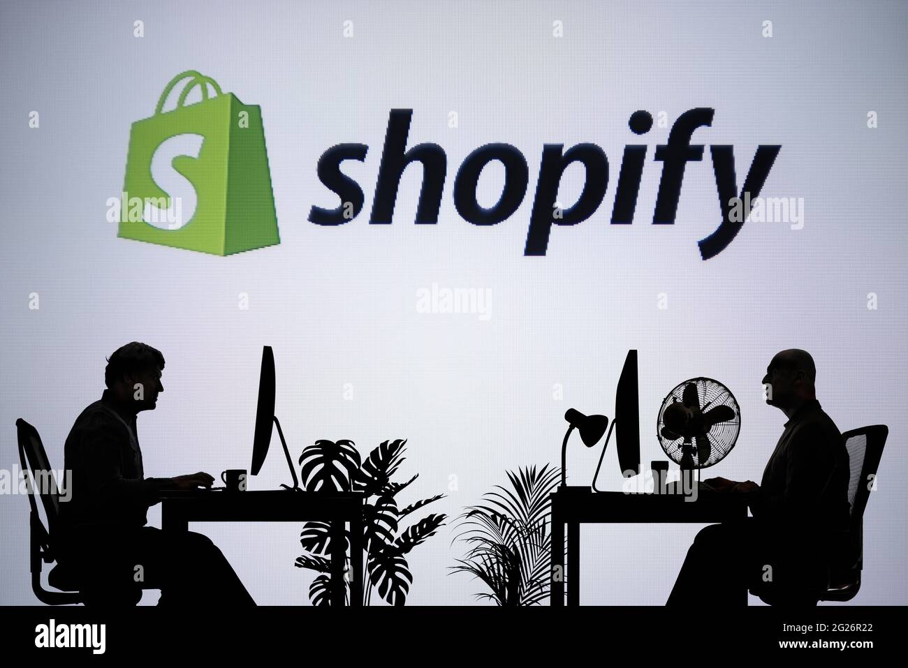 Le logo Shopify est visible sur un écran LED en arrière-plan tandis que deux personnes silhouetées travaillent dans un environnement de bureau (usage éditorial uniquement) Banque D'Images