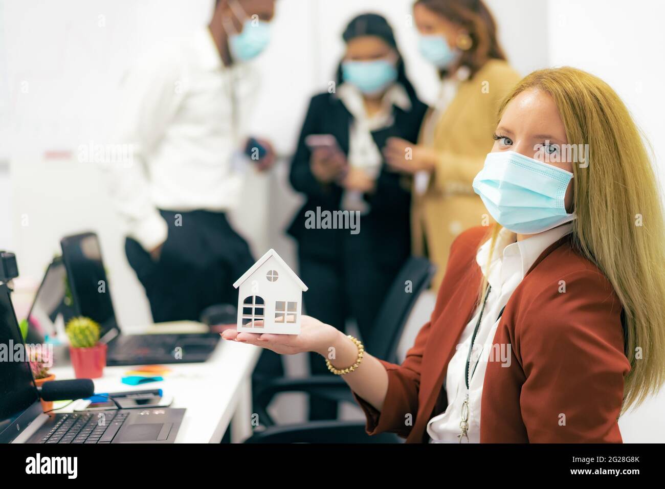 Jeune femme avec masque de protection montrant la maison blanche au bureau. Agent immobilier et concept de coronavirus. Banque D'Images