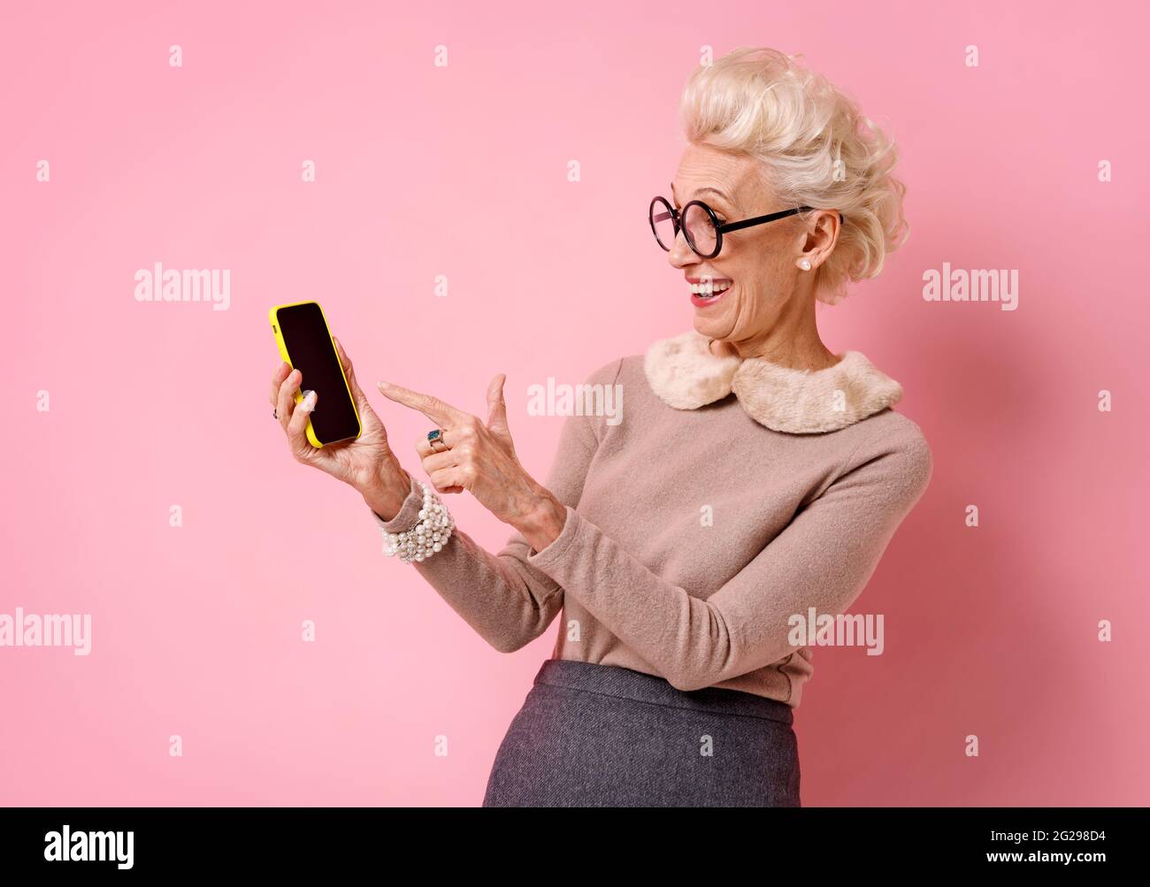 Une grand-mère souriante pointe le doigt sur l'écran du téléphone. Photo d'une femme âgée sur fond rose. Banque D'Images