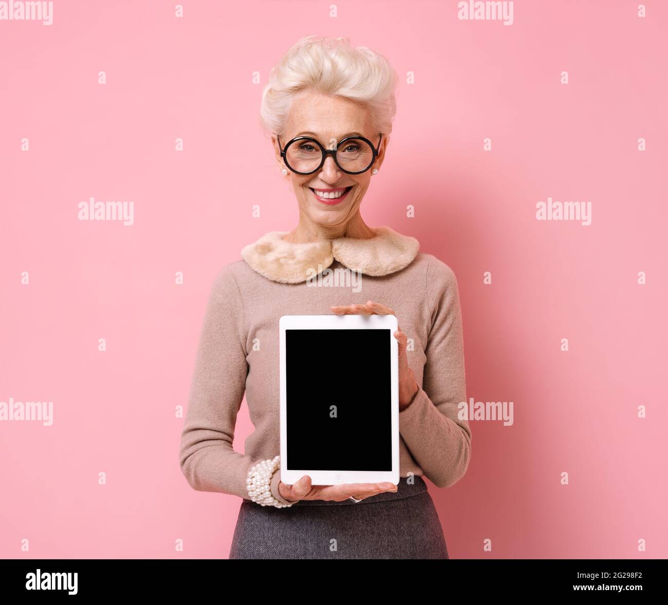 Grand-mère tient une tablette numérique avec un écran vide. Photo d'une femme âgée sur fond rose. Banque D'Images
