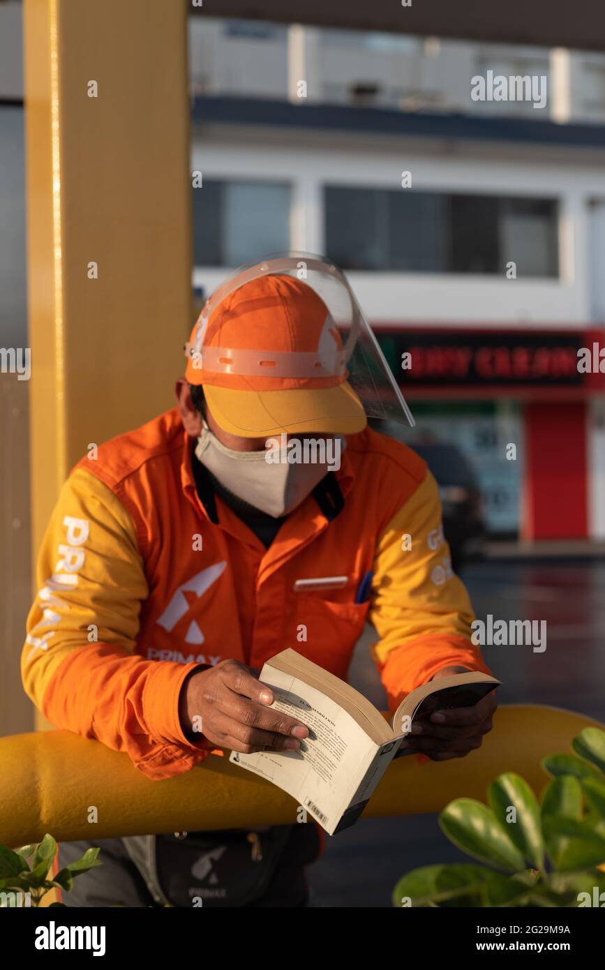 Jeune ouvrier péruvien portant un uniforme orange et un masque Covid lit le livre Silmarillion dans la station-service Primax où il travaille à Lima. Banque D'Images