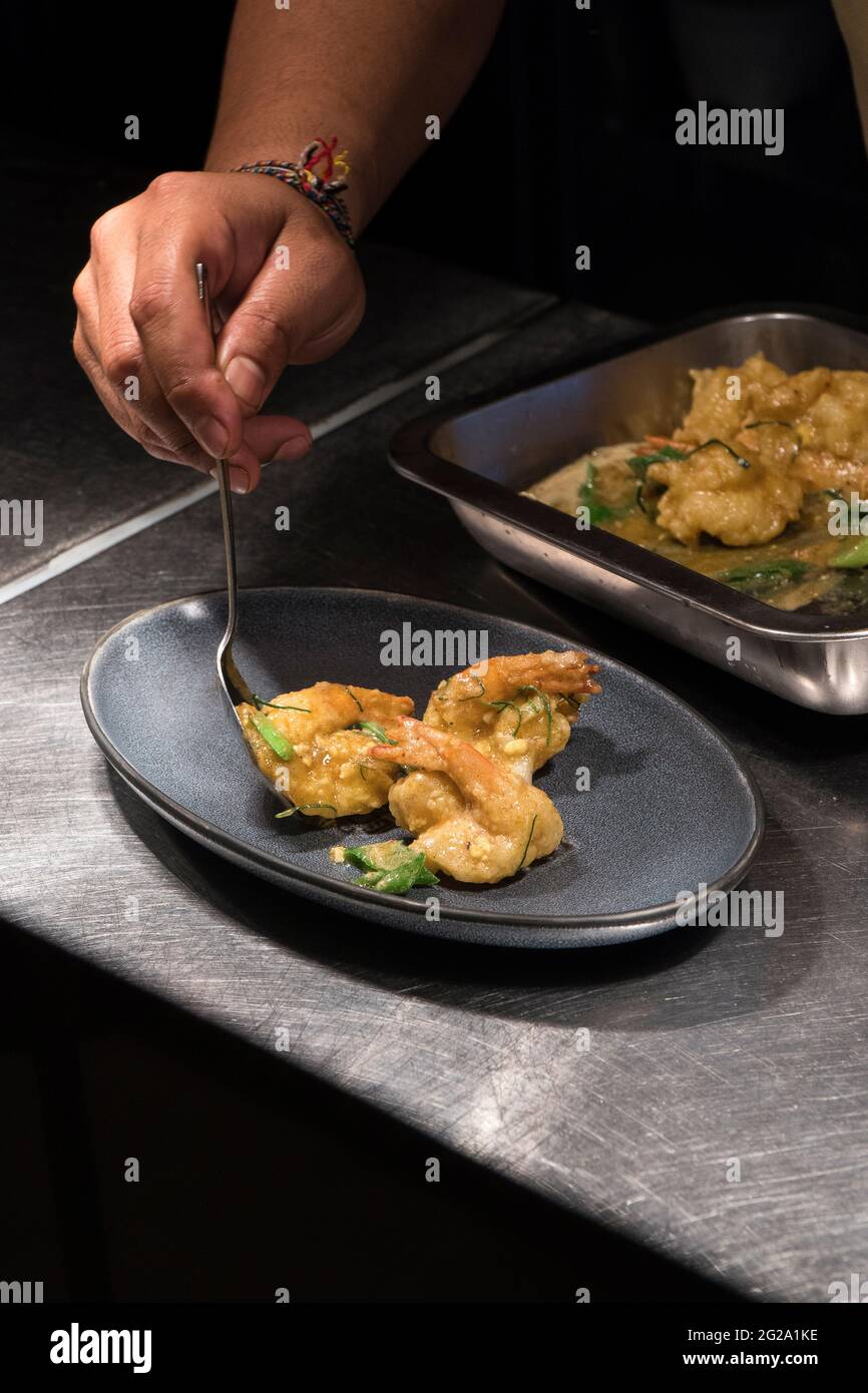 Chef cuisinier haut de gamme servant des crevettes frites avec des herbes fraîches vertes sur une plaque ovale grise tout en se tenant sur un comptoir métallique dans un cadre contemporain Banque D'Images
