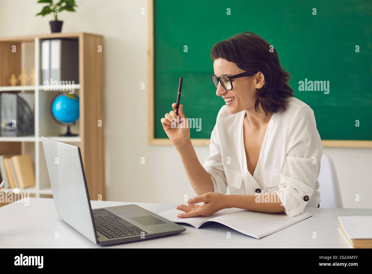 Bon enseignant assis au bureau avec un ordinateur portable et ayant un cours virtuel avec des élèves Banque D'Images