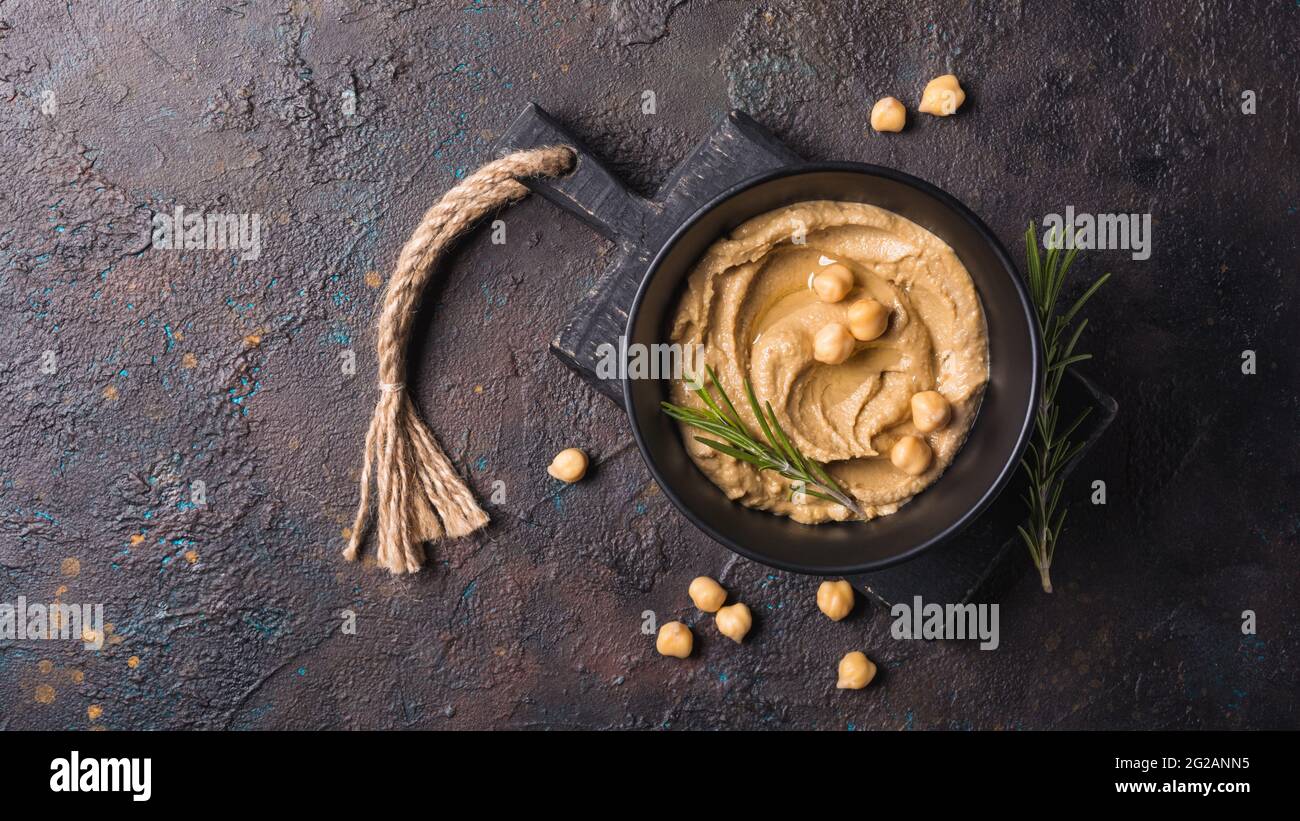 Vue de dessus de l'en-cas de houmous de pois chiches maison traditionnel avec épice de romarin vert frais dans un bol noir sur fond de béton foncé Banque D'Images