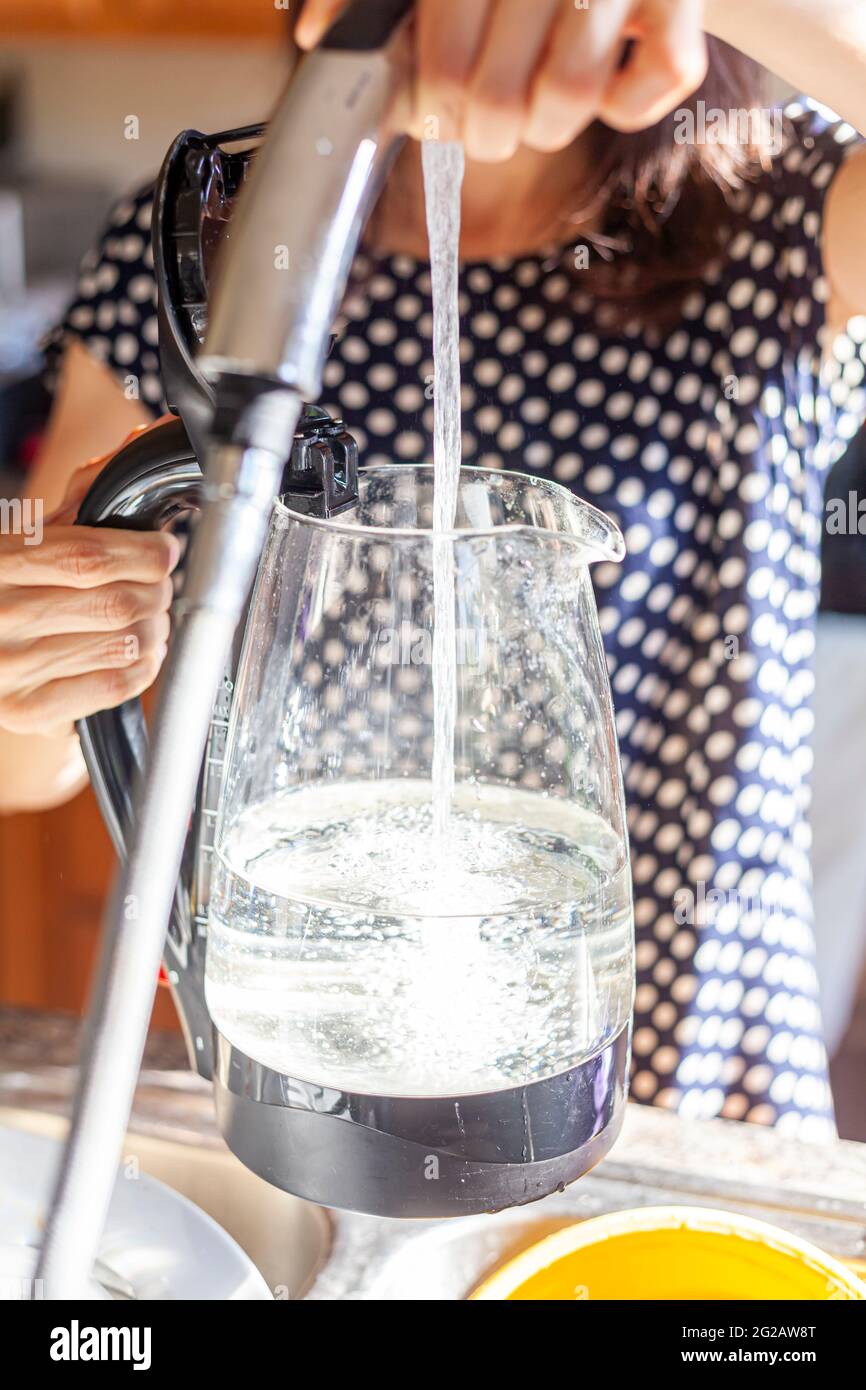 Une femme portant une robe d'été à motif à pois remplit une bouilloire électrique en verre avec le raccord pulvérisateur du robinet de cuisine. Elle étend le Banque D'Images
