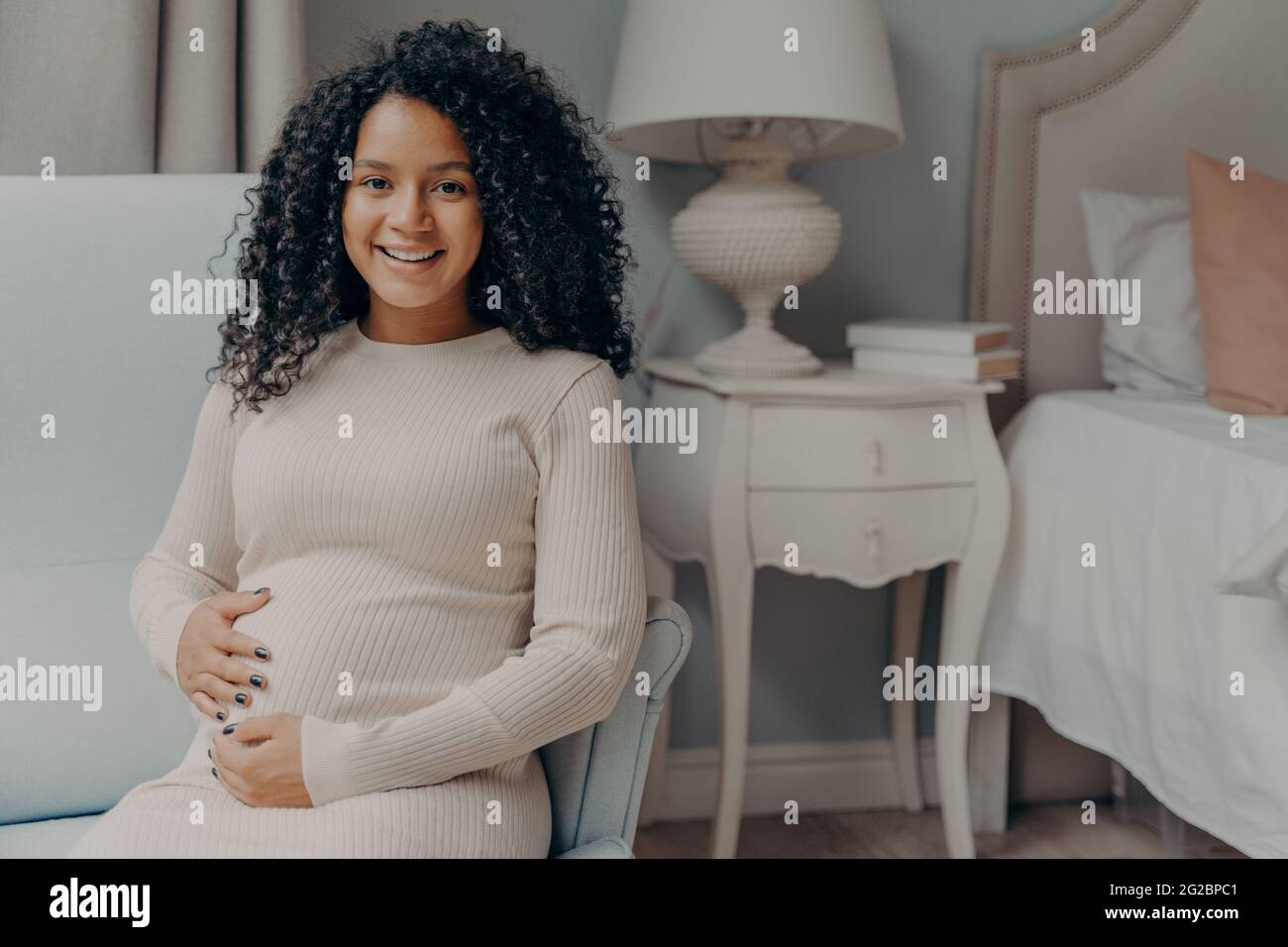 Jeune femme enceinte heureuse avec des cheveux bouclés tenant son ventre et  souriant à la caméra Photo Stock - Alamy