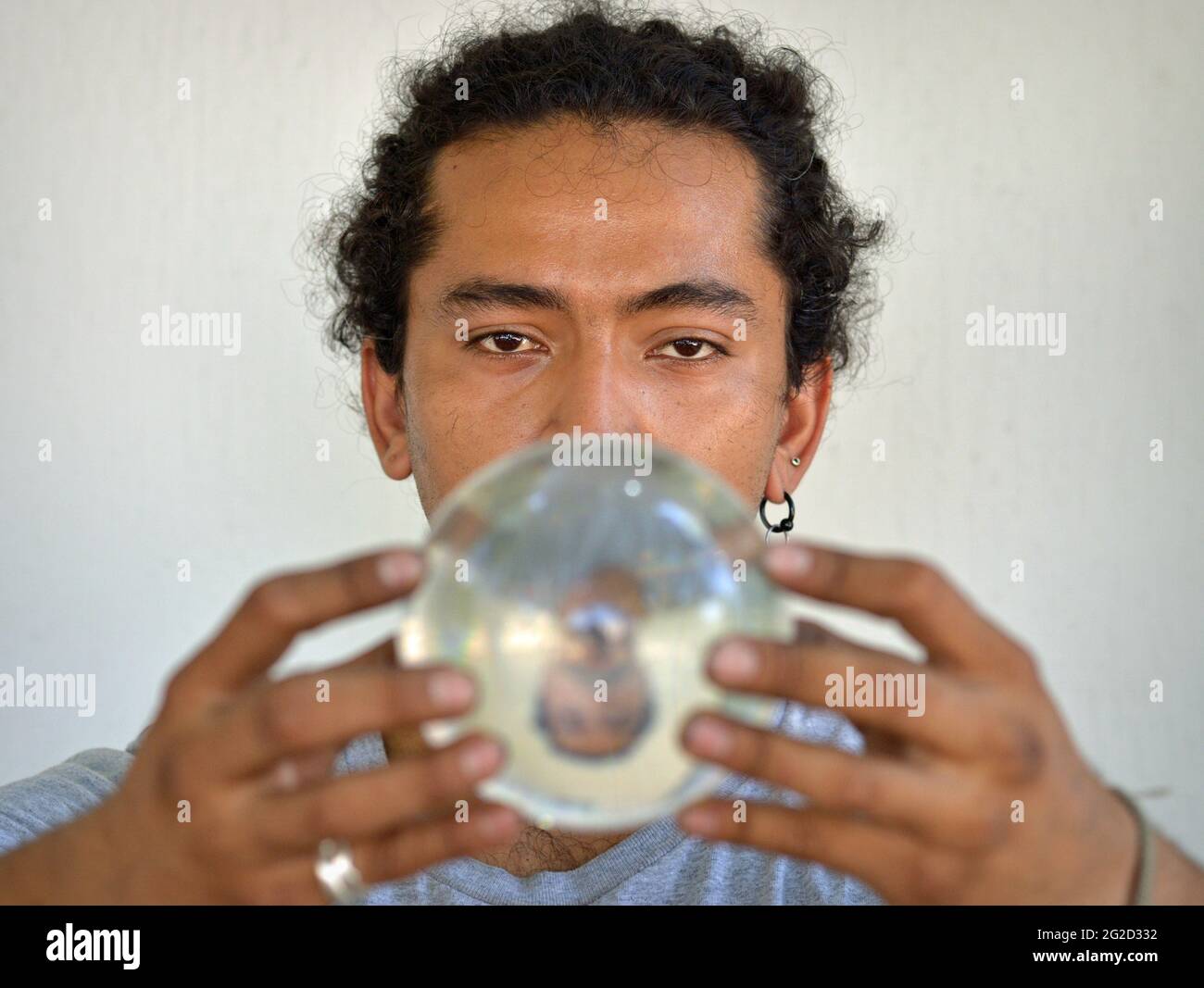 Beau jeune Latino-homme avec des boucles tient une boule de cristal avec son image miroir à retournement vertical et fixe au spectateur avec des yeux fascinants. Banque D'Images