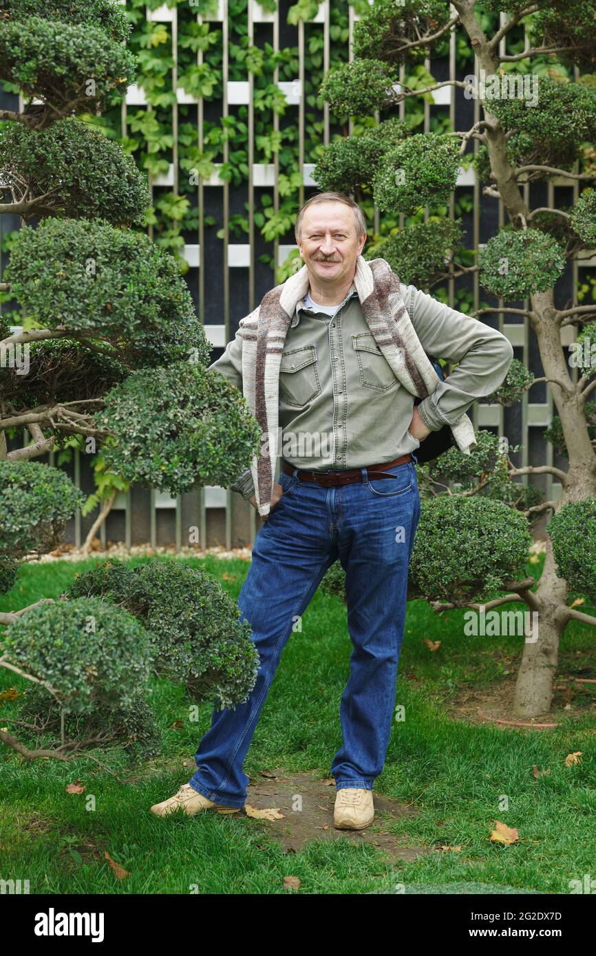 Un homme de cinquante ans aux cheveux blancs en Jean et une chemise se tient dans le parc de la ville près d'un arbre et regarde l'appareil photo avec le sourire Banque D'Images