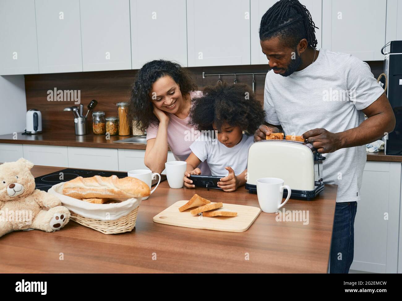 Le père, la mère et la petite fille préparent la nourriture dans la cuisine de leur maison. Recette de plat de recherche d'enfant sur Internet en utilisant un smartphone pour sa famille Banque D'Images