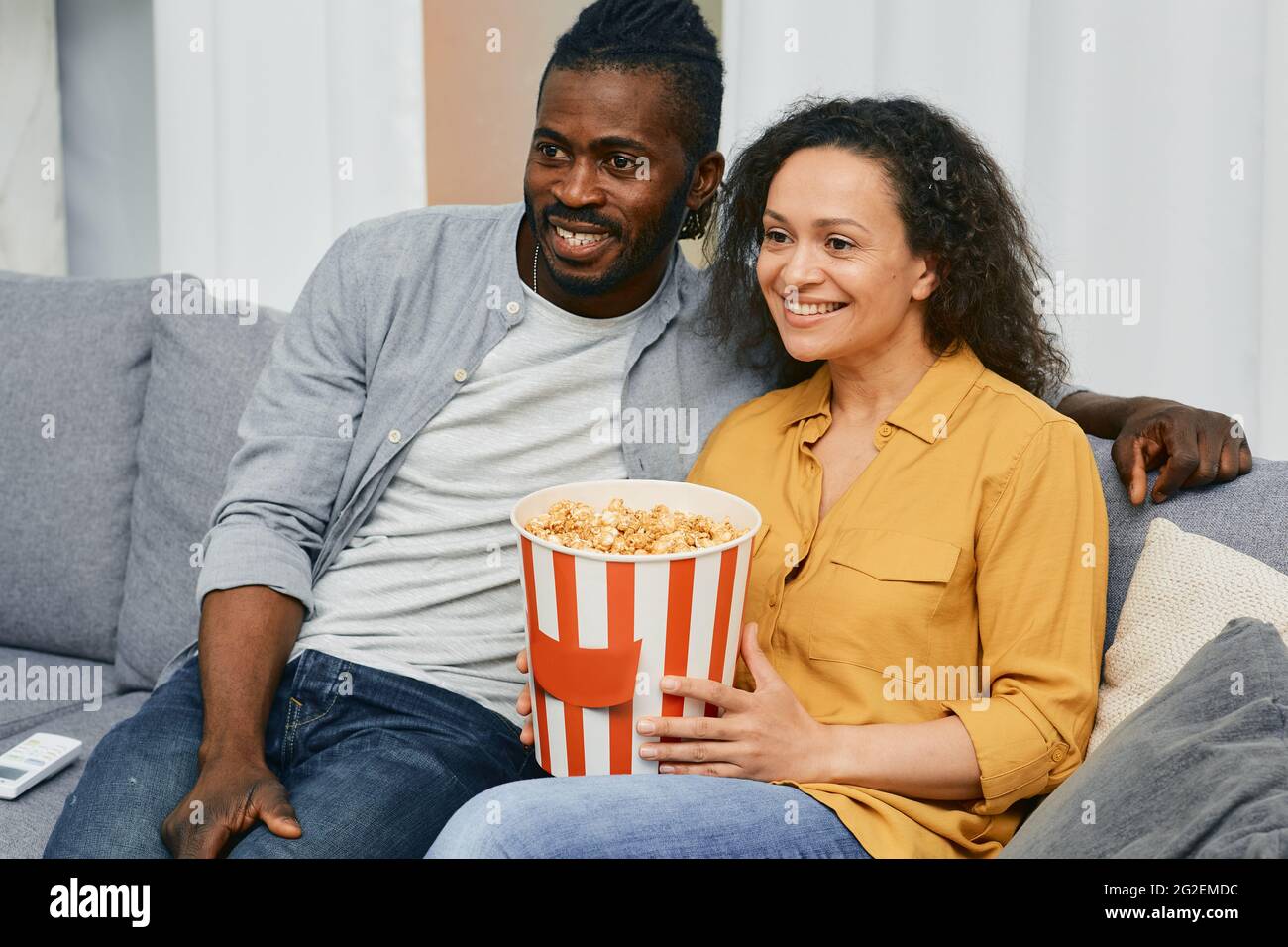 Couple de course mixte regardant la télévision avec un grand seau de pop-corn dans leur maison tout en étant assis sur un canapé Banque D'Images