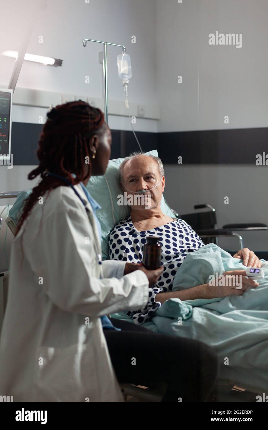 Thérapeute afro-américain dans la chambre d'hôpital discutant du traitement tenant des pilules bouteille rassurant de récupération, malade homme âgé allongé dans le lit respiration par le masque d'oxygène, Banque D'Images