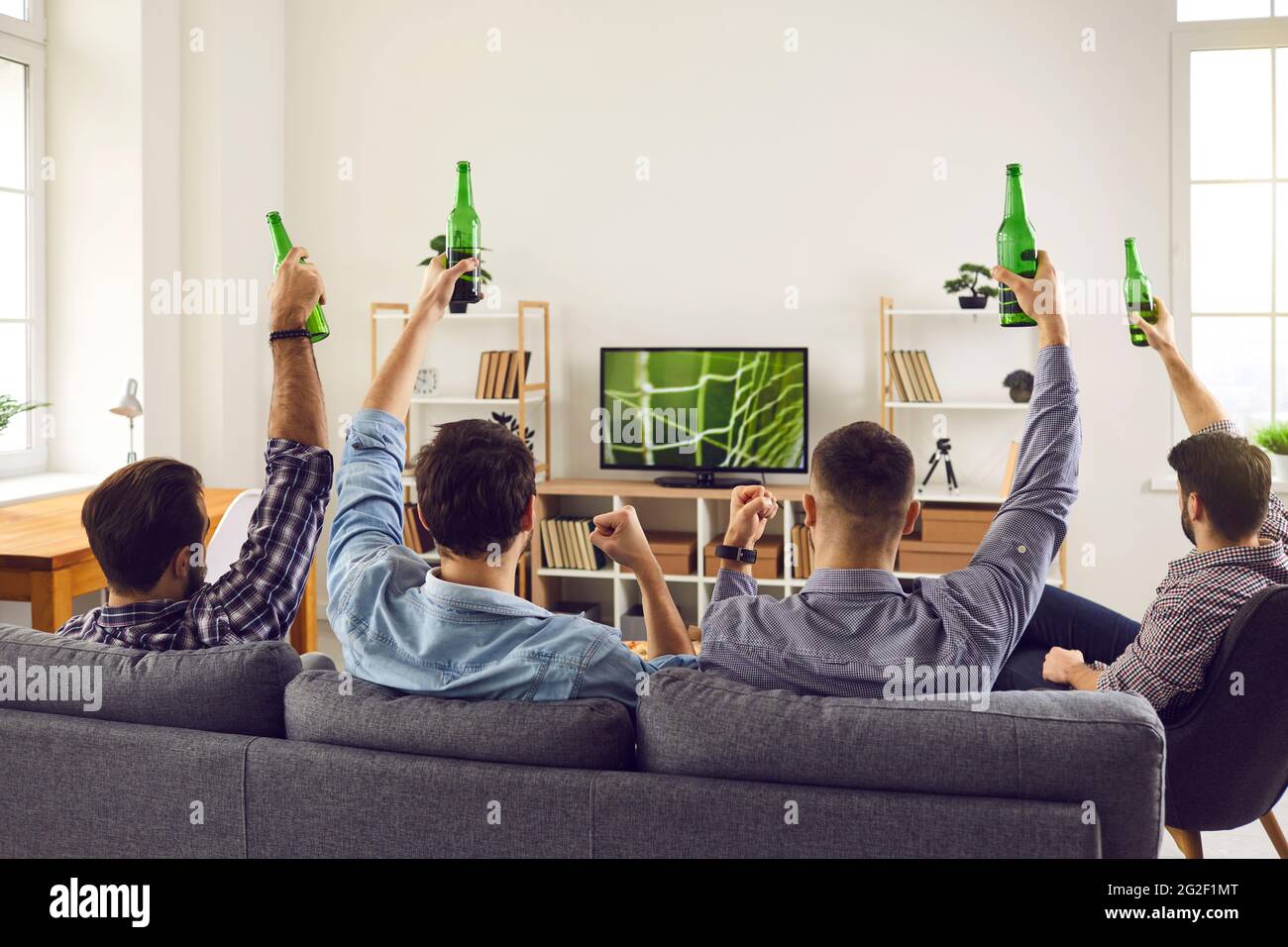 Un groupe de jeunes amis regardent un match de football à la maison à la télévision, tenant une bouteille Banque D'Images