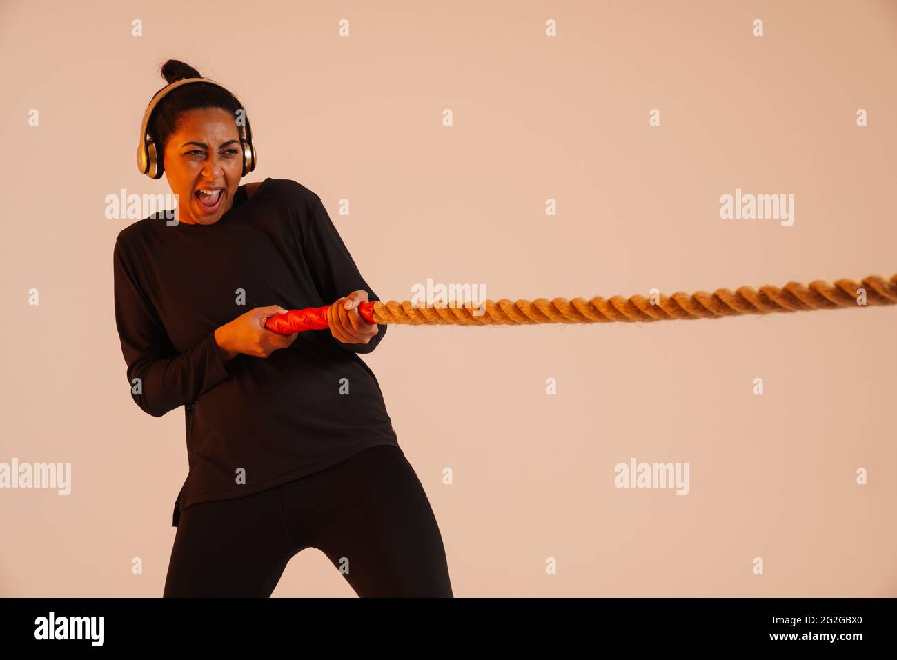 Femme afro-américaine athlétique dans un casque d'écoute s'exécutant avec corde de bataille isolée sur fond beige Banque D'Images