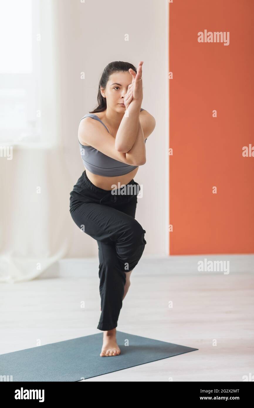 Jeune femme entraîneur pratiquant le yoga, debout sur le tapis en studio, exécute l'exercice de garudasana, la pose d'aigle. Banque D'Images