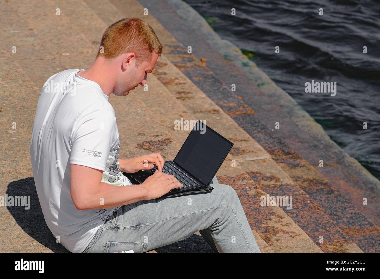 Un jeune homme travaille sur un ordinateur assis sur le remblai de la rivière Neva près de l'eau sur les marches de granit. Saint-Pétersbourg. Russie. Juin 9 2021 Banque D'Images