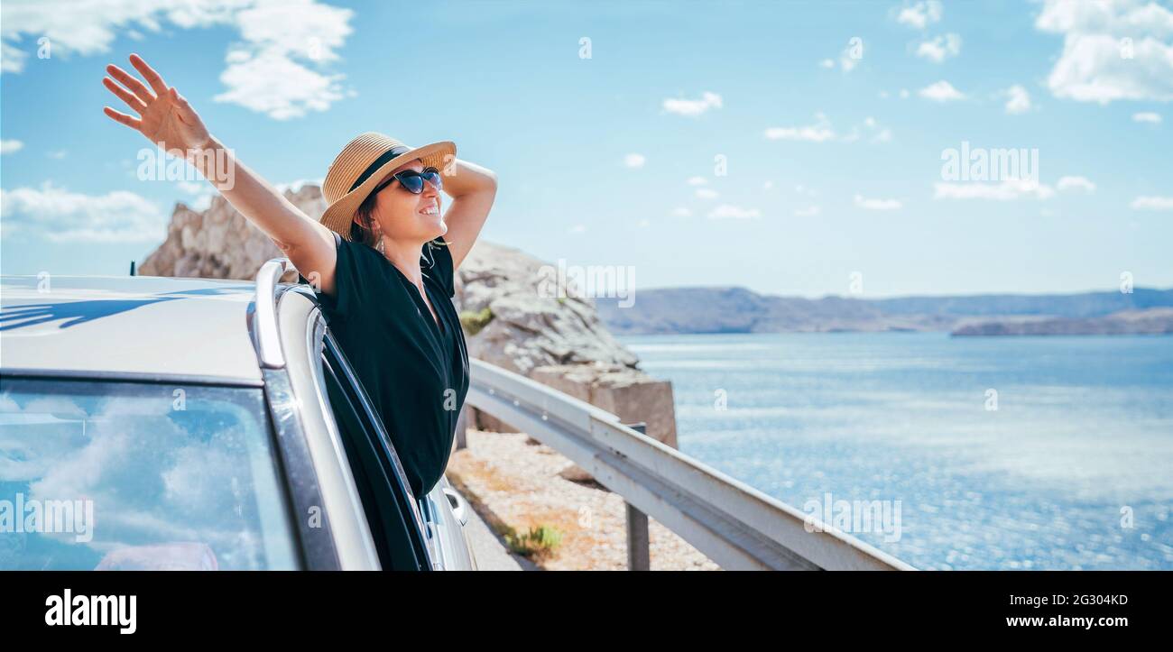 Portrait de femme joyeuse appréciant le voyage en bord de mer. Vêtue d'une robe noire, chapeau de paille et lunettes de soleil, elle ouvrit les bras et brillant avec le happine Banque D'Images