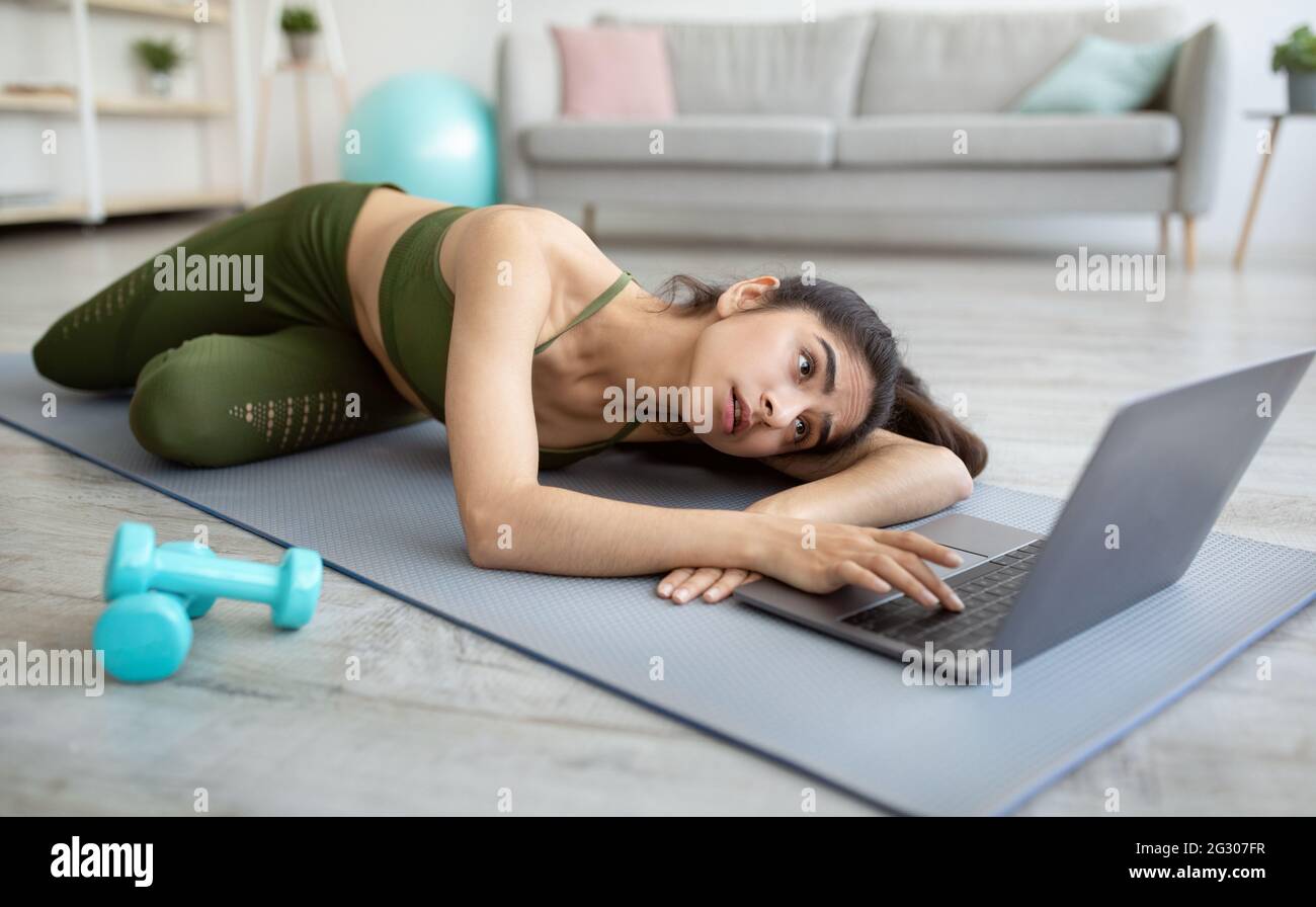 Sports en ligne. Une jeune femme indienne fatiguée, allongé sur un tapis de yoga près d'un ordinateur portable, n'ayant aucune force pour l'entraînement domestique Banque D'Images