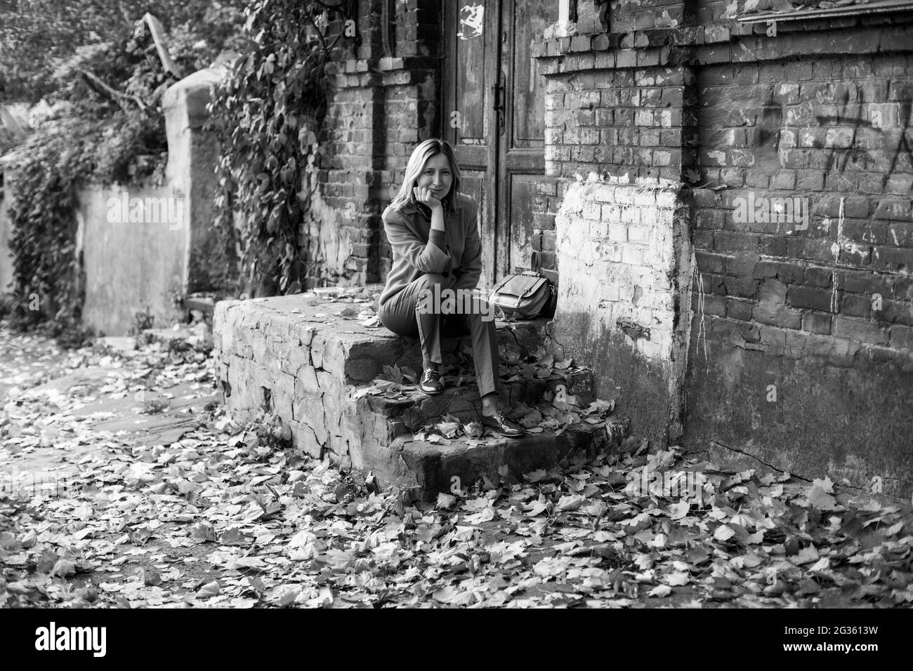 Une femme élégante dans une veste est assise sur le porche d'une vieille maison avec des feuilles d'érable sur le sol dans l'atmosphère de l'automne. Photo en noir et blanc. Banque D'Images