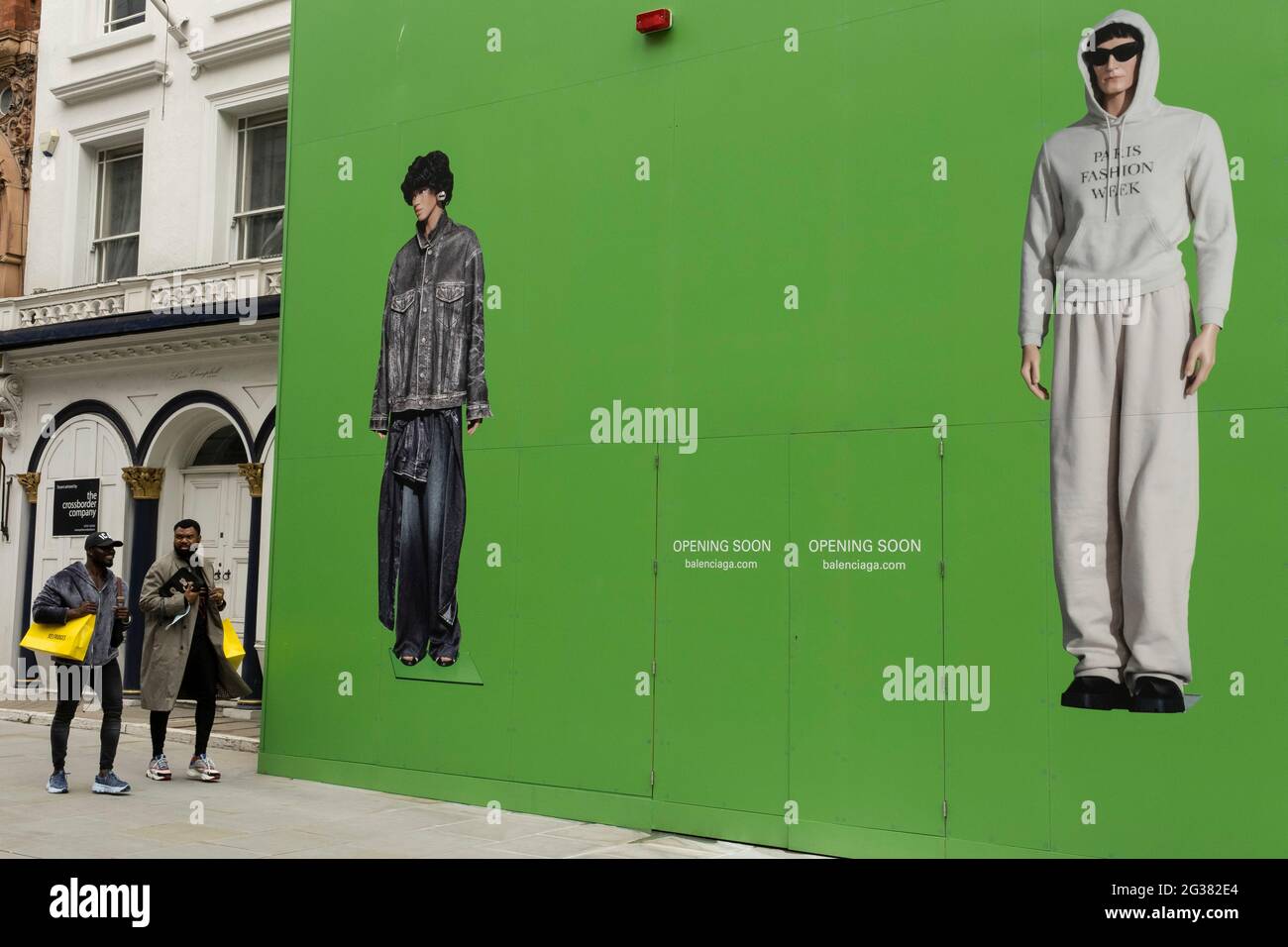 Les gens interagissent avec des personnages d'action à grande échelle  portant des vêtements tendance contre une énorme palissade verte qui couvre  le magasin Balenciaga lors d'une remise en place dans la zone