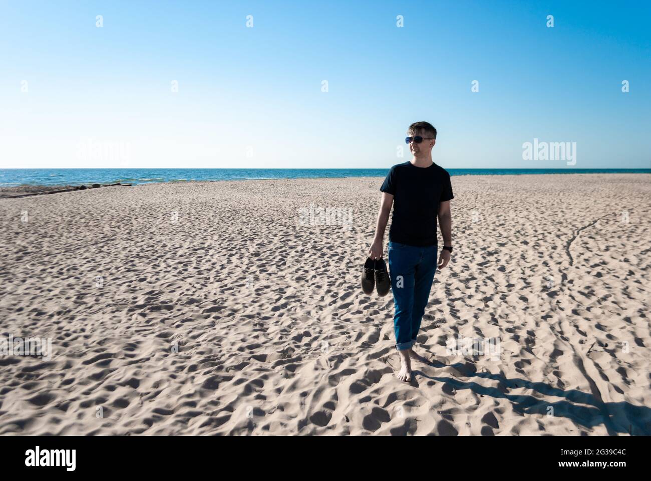 Homme en tenue décontractée, lunettes se tient sur la plage et tient des chaussures à la main Banque D'Images