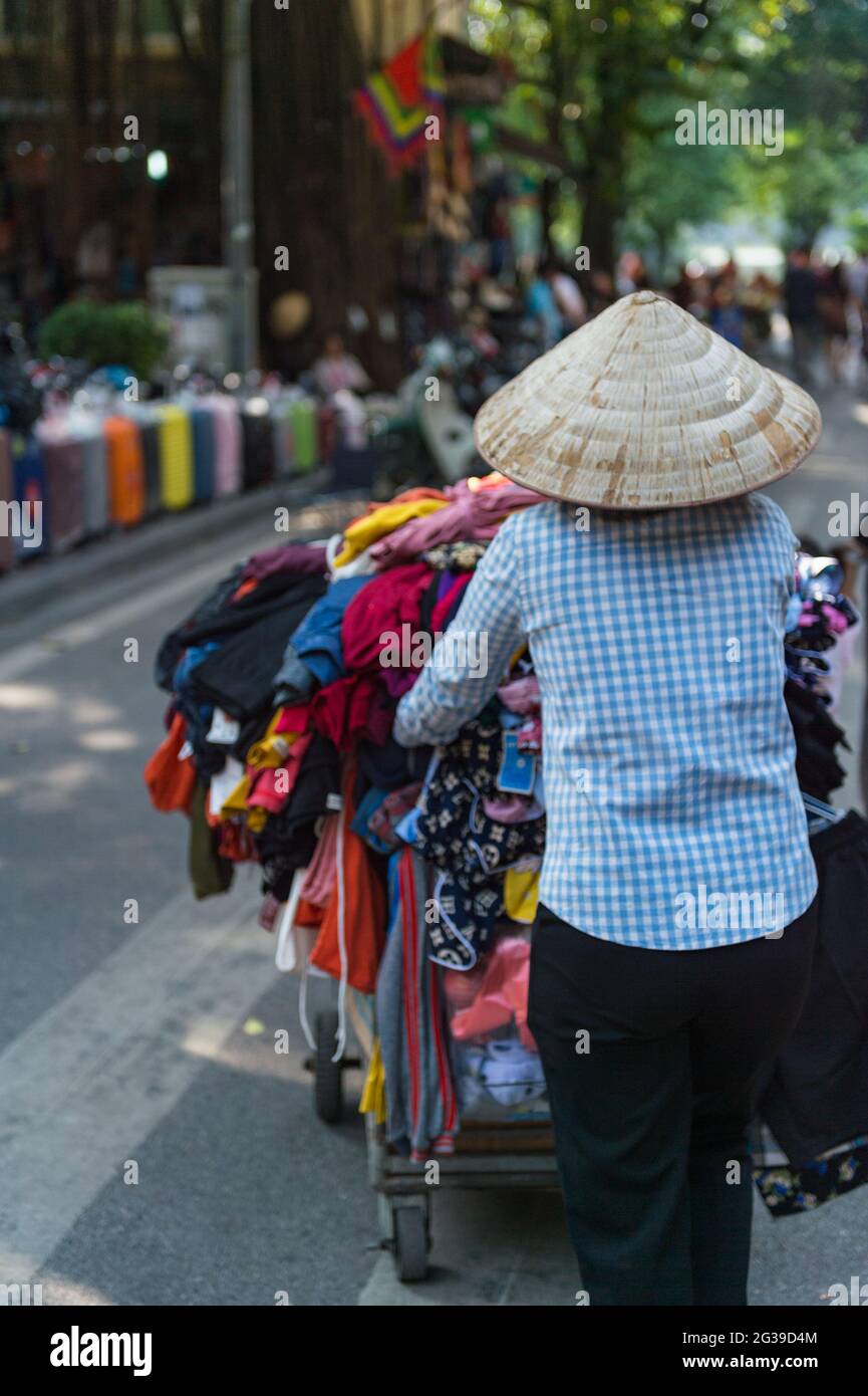 Un marchand de rue portant un chapeau poussant un chariot plein de vêtements à Hanoi, Vietnam Banque D'Images