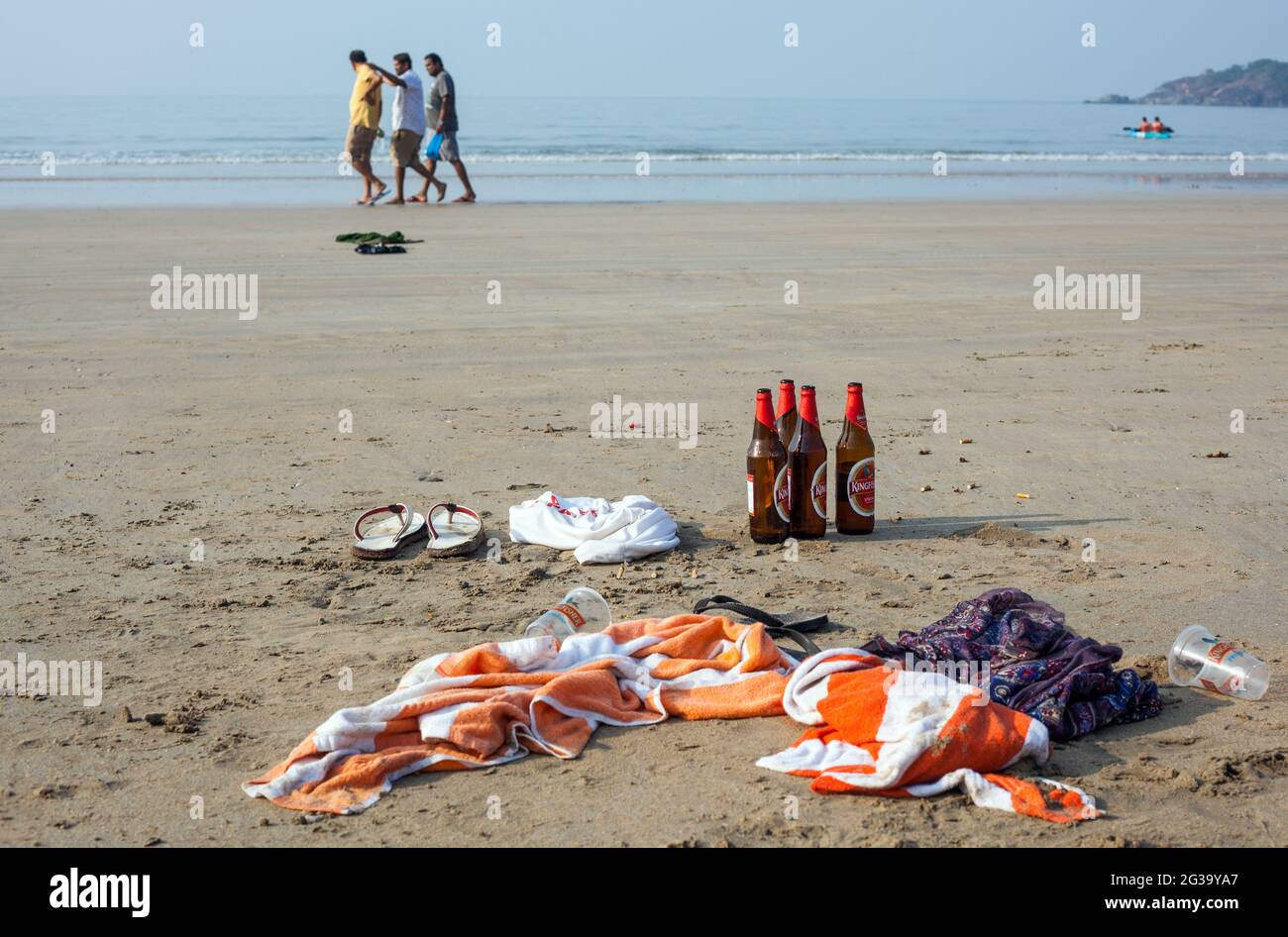 Bouteilles de bière et serviettes laissées sur une plage vide et trois gars indiens se promènent au bord de la mer, Agonda, Goa, Inde Banque D'Images