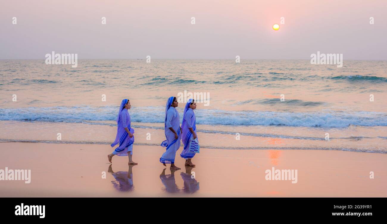 Vue panoramique de trois religieuses catholiques marchant le long de la plage pieds nus dans le sable humide au coucher du soleil, Agonda, Goa, Inde Banque D'Images