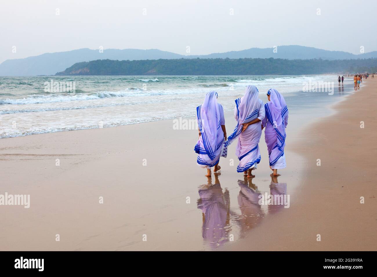 Vue arrière de trois religieuses catholiques marchant le long de la plage pieds nus dans le sable humide, Agonda, Goa, Inde Banque D'Images