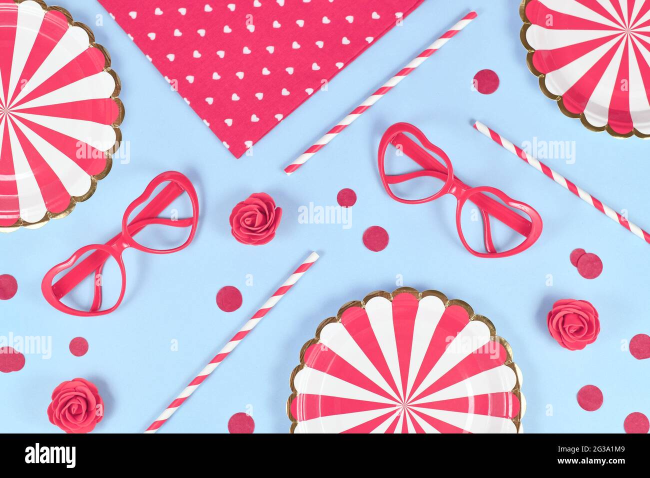 Plat de fête avec assiettes en papier, verres en forme de coeur, roses, confetti et pailles à boire sur fond bleu Banque D'Images