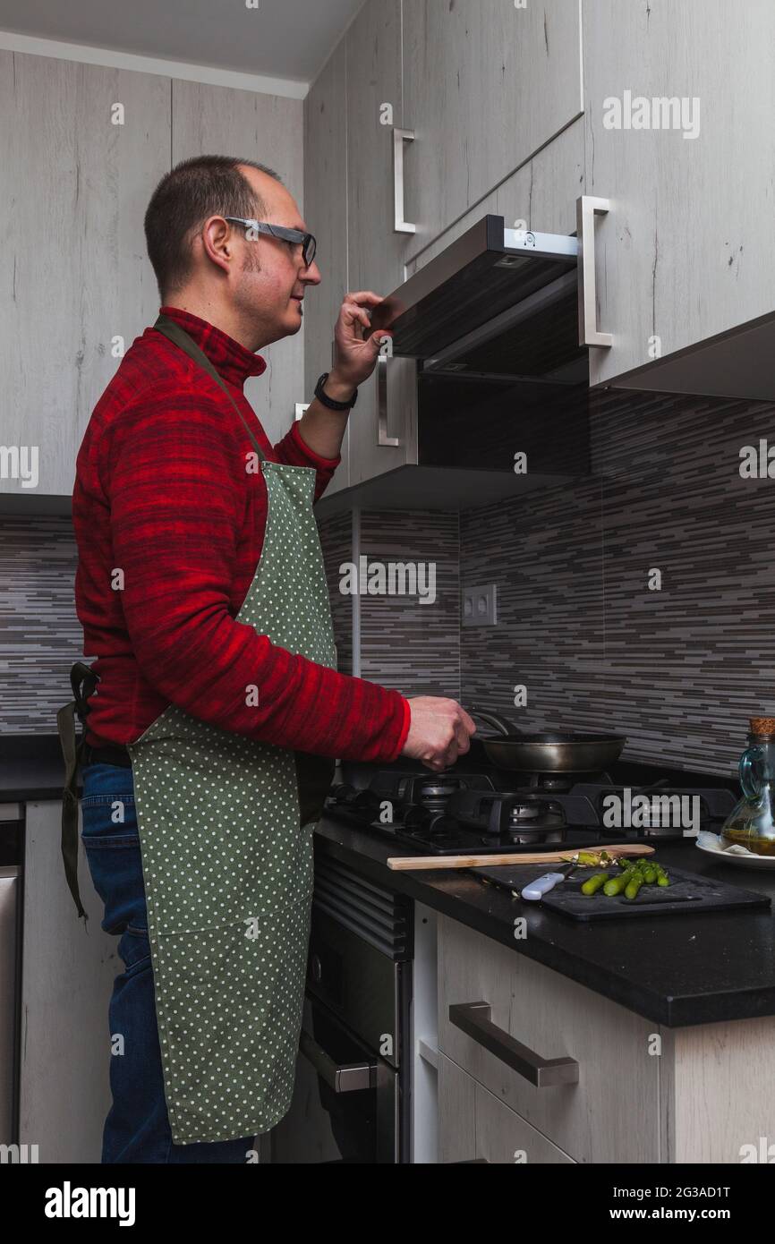 Un homme en polaire rouge et un tablier vert cuisant des artichauts grillés et des asperges dans une cuisine. Il ouvre le capot de l'extracteur. Banque D'Images