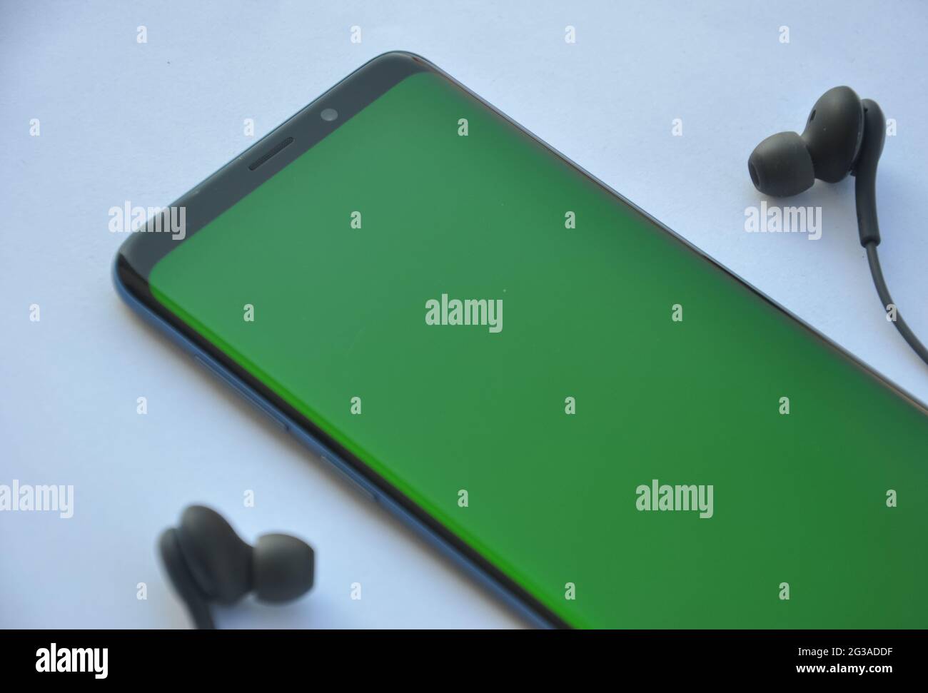 Vue à faible angle d'un téléphone portable à écran incurvé avec écran vert et écouteurs noirs sur fond blanc Banque D'Images