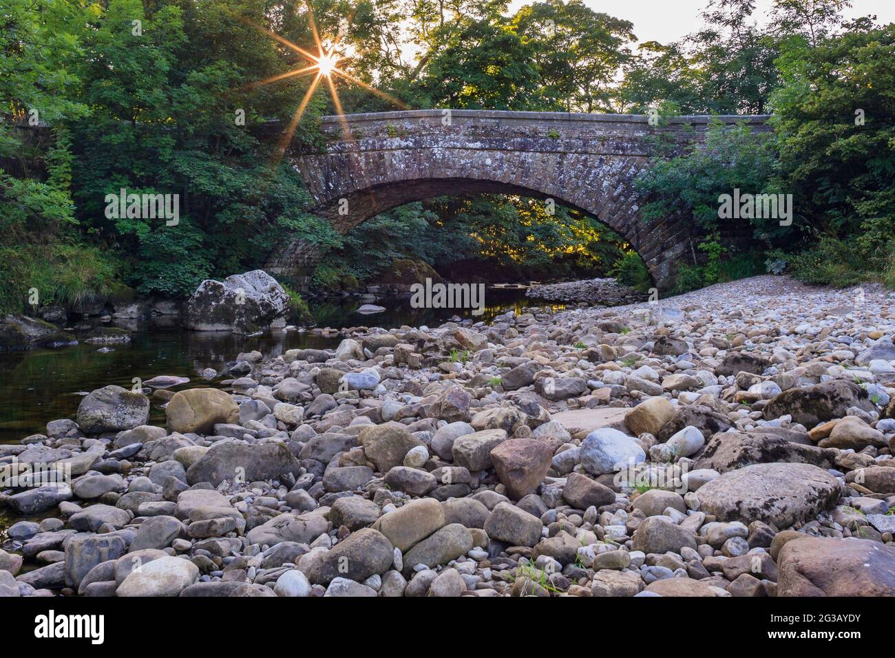 Sunburst raies sur le vieux pont en pierre (1 arche) et la rive rocheuse par les eaux peu profondes de la rivière - pittoresque Wharfe Valley, Hubberholme, Yorkshire Dales, Angleterre, Royaume-Uni. Banque D'Images