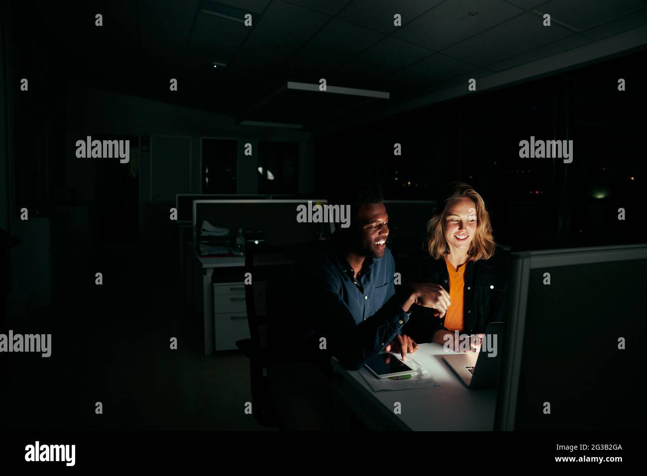 Deux collègues heureux et divers travaillant sur un ordinateur portable jusqu'à la nuit dans un bureau sombre Banque D'Images