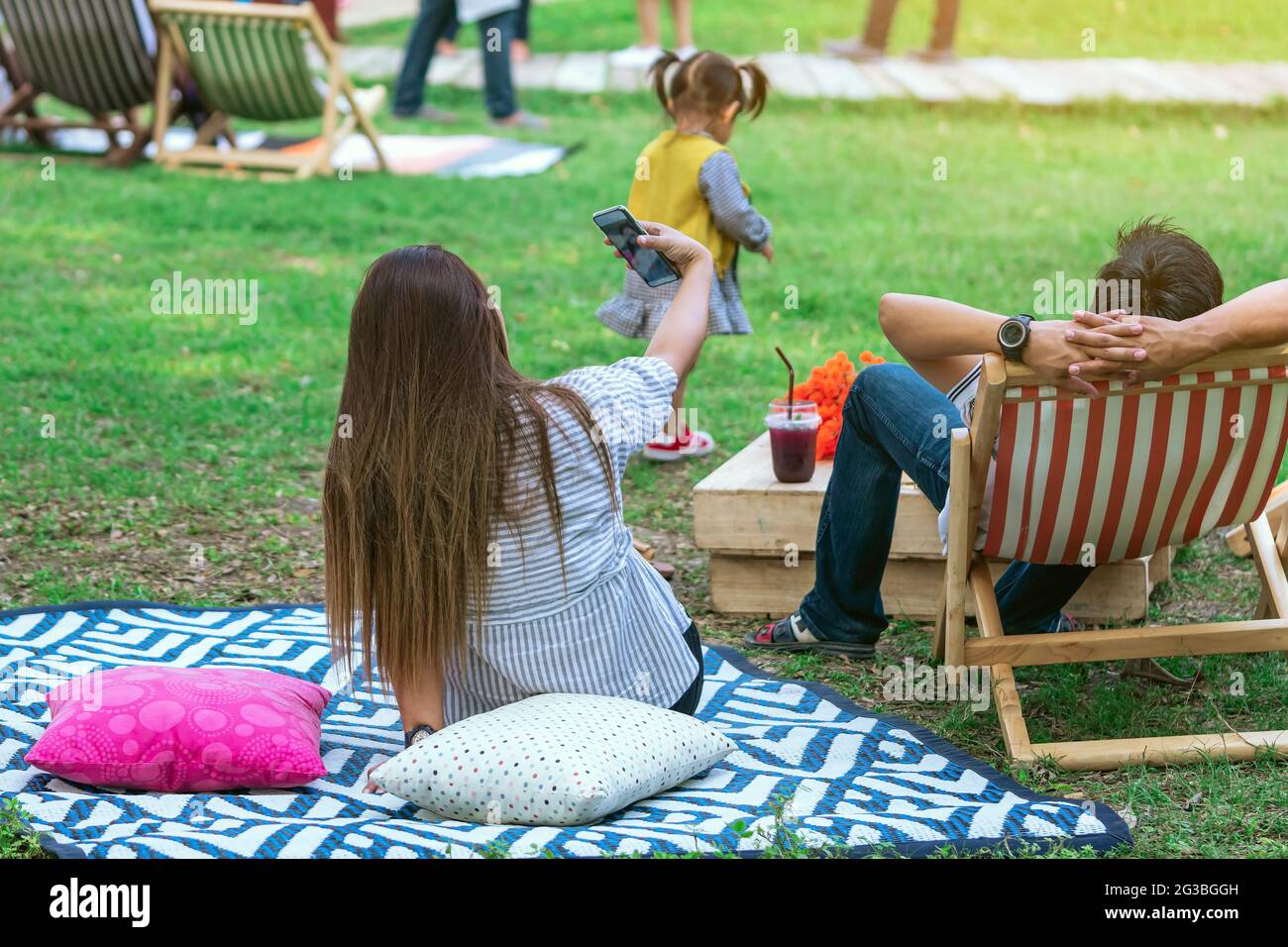Vue arrière d'une femme asiatique s'assoir pour prendre le selfie sur le  tapis près de son amant sur la chaise de pont dans le jardin. Vacances  d'été dans un cadre verdoyant. Personne