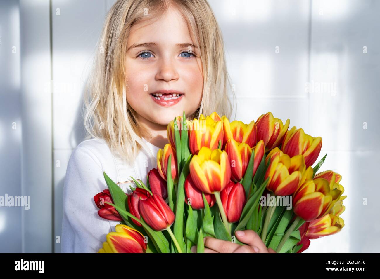 petite fille sans dents souriante avec un énorme bouquet de tulipes sur fond blanc avec lumière directe du soleil et ombres. Obtenir des fleurs. Bouquet cadeau Banque D'Images