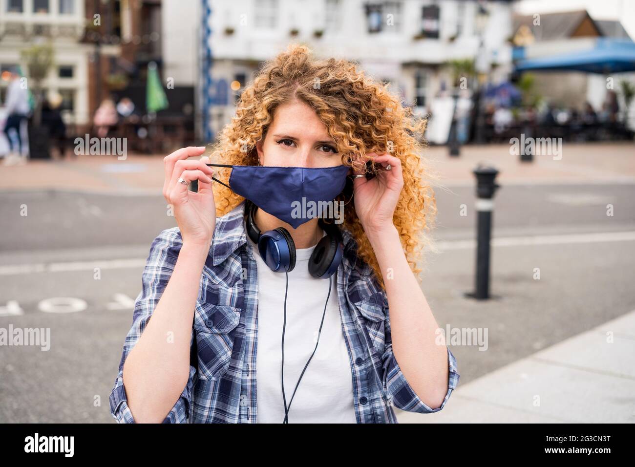 Une jeune femme caucasienne heureuse prend un masque protecteur couvrant le visage à l'extérieur sur fond de ville européenne. La pandémie Covid-19 est terminée Banque D'Images