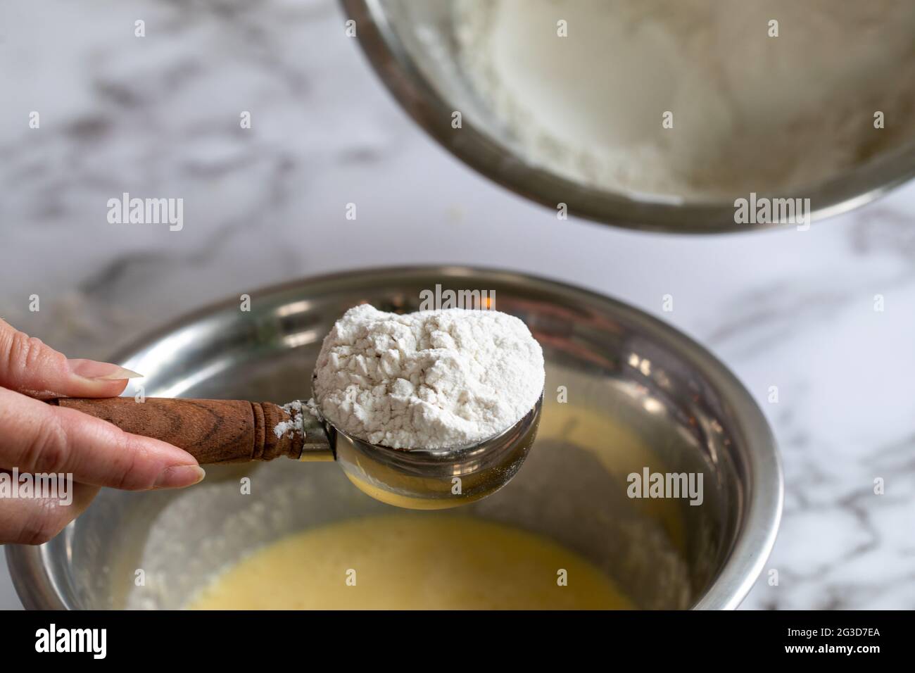 Main de femme tenant une cuillère de mesure pleine de farine sur un saladier rond en métal rempli d'un mélange jaune pour la pâtisserie Banque D'Images