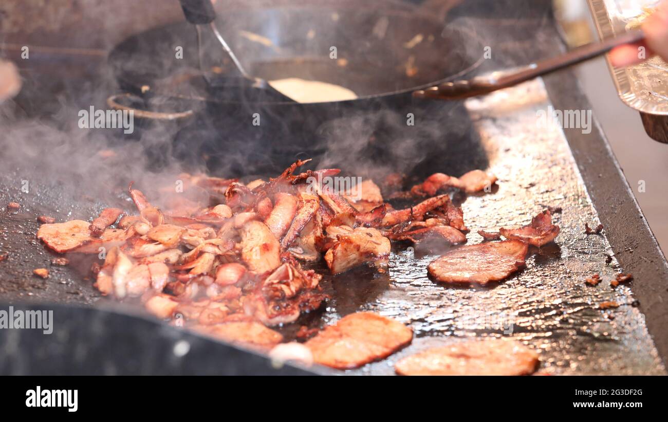 Gros plan de beaucoup de bacon croustillant cuit sur une plaque de barbecue chaude avec une cuisson à la crêpe dans une casserole en arrière-plan. Banque D'Images