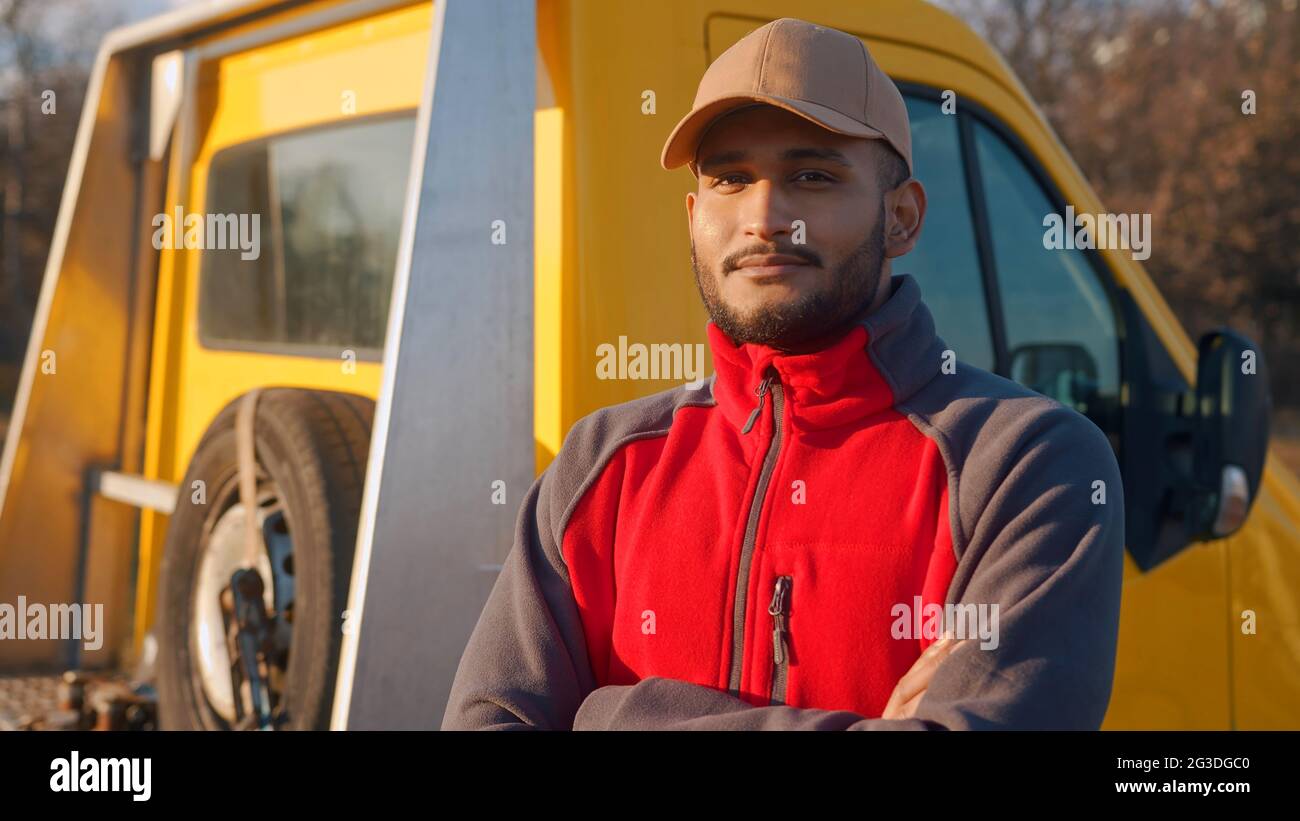 Un employé portant un chapeau et un uniforme avec un camion jaune derrière lui. Garçon souriant à la caméra et debout avec les bras croisés. Concept de mécanicien automobile. La roue de secours est visible dans le chariot derrière. Banque D'Images
