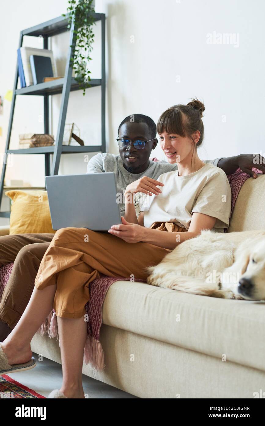 Un jeune couple multiethnique assis sur un canapé regardant quelque chose sur un ordinateur portable et souriant qu'il passe du temps à la maison Banque D'Images