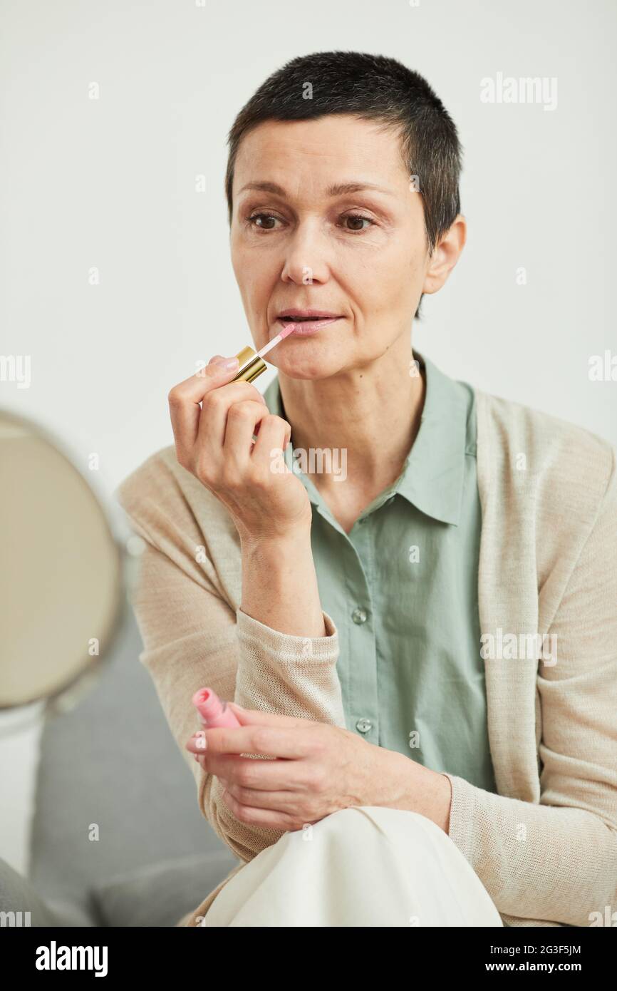 Portrait vertical franc de la femme mûre mettant sur le rouge à lèvres tout en faisant le maquillage à la maison et regardant dans le miroir Banque D'Images