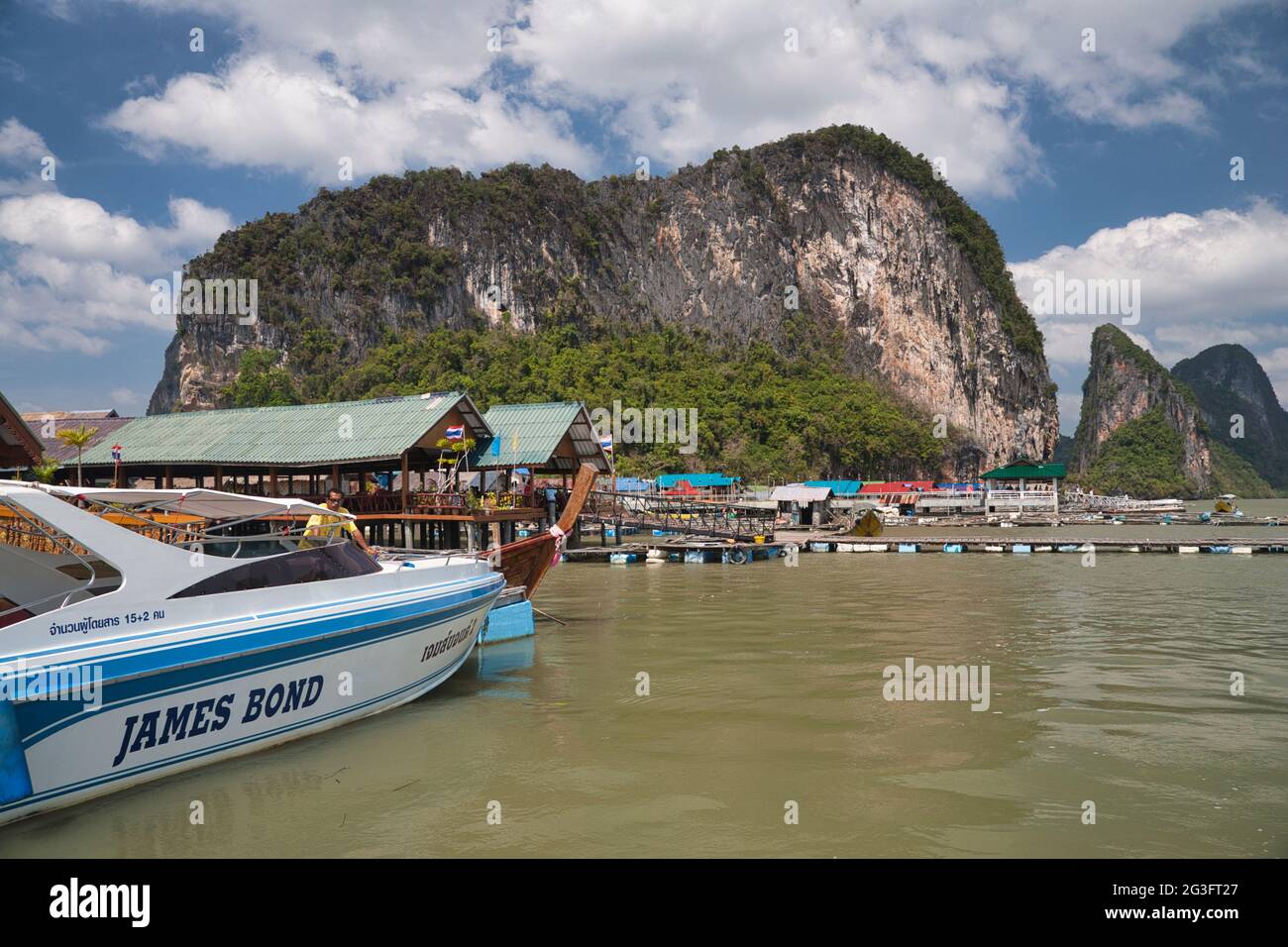 Un bateau à moteur moderne amarré dans un village aquatique construit sur pilotis à Phang Nga, en Thaïlande, avec d'énormes rochers et falaises en arrière-plan Banque D'Images