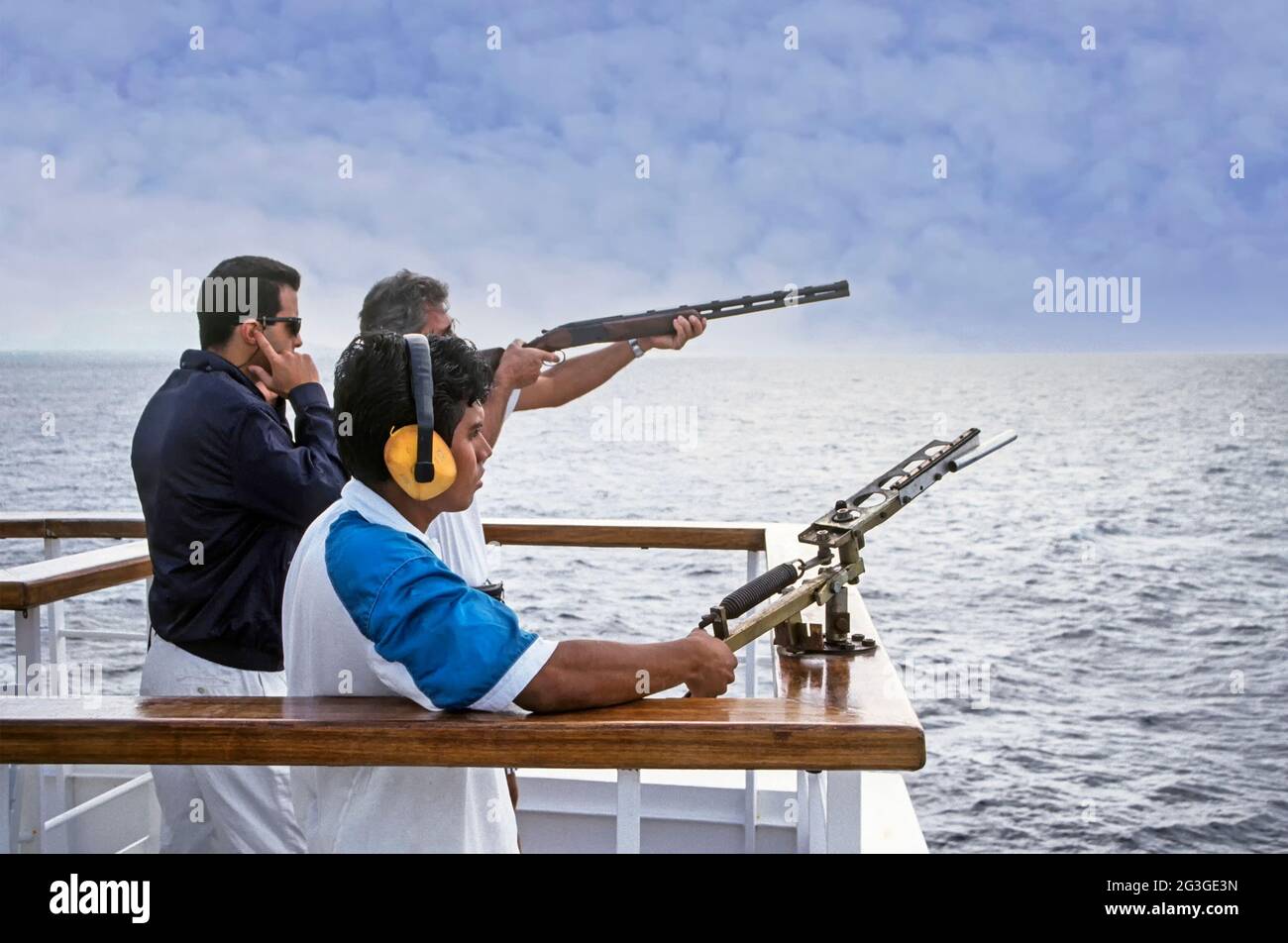 Archive historique des années 90 image de gros plan de tir de pigeon d'argile ou d'argile cible une activité sportive de tir avec l'équipement fourni pour les voyageurs de croisière sur la poupe d'un navire de croisière des années 1990, paquebot en mer utilisé avec une machine boulonnée à la main courante qui propulse le piège cible au-dessus de l'eau de mer tiré par la main de pont portant des défenseurs d'oreilles et d'homme passager visant une arme à feu de fusil supervisée par un agent de divertissement de navires avec les doigts au-dessus des oreilles une image d'archives du chemin Nous avons profité de vacances en croisière dans la mer Méditerranée en 1993 Banque D'Images