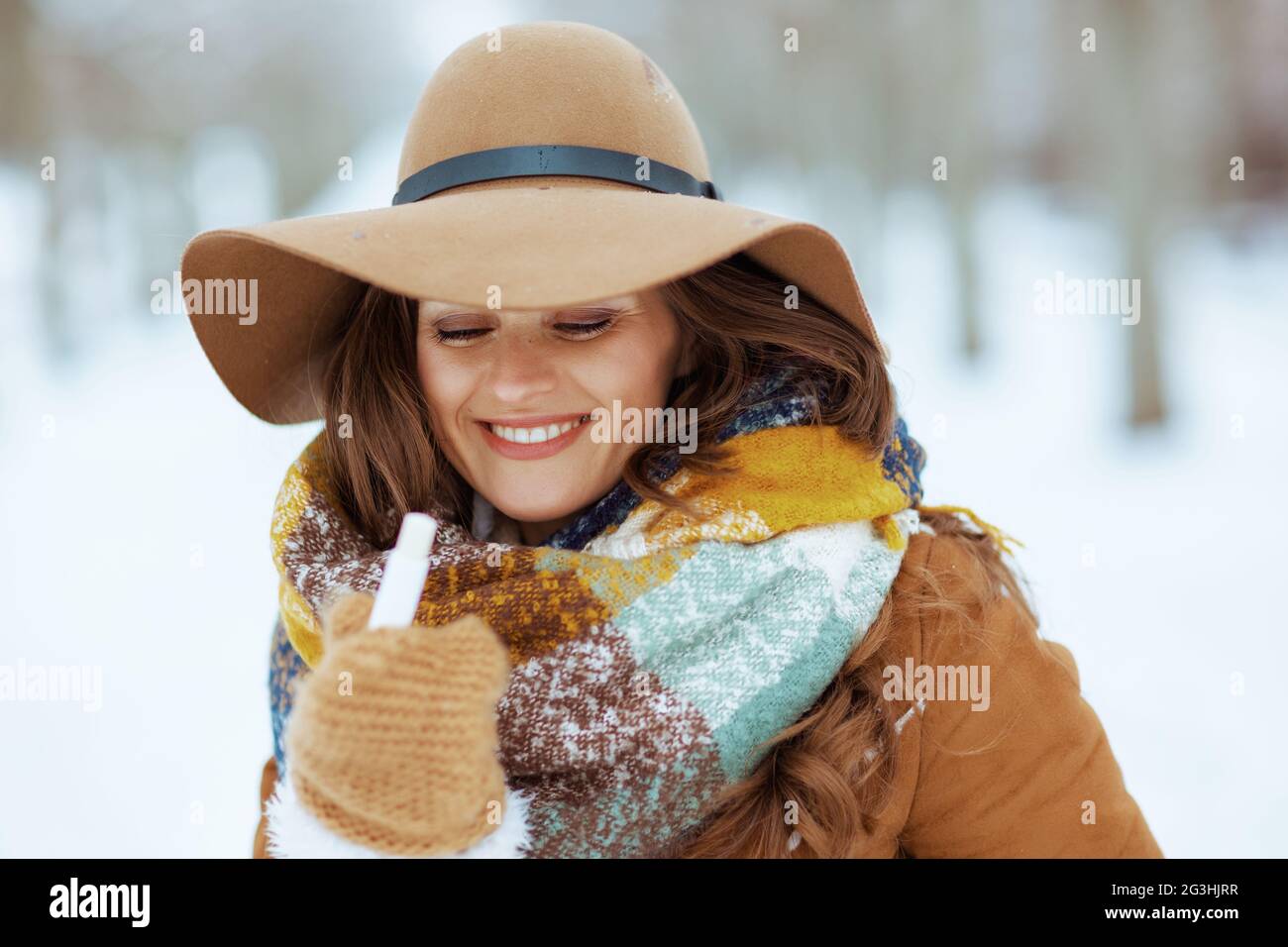 bonne femme élégante de 40 ans en chapeau marron et écharpe avec moufles et  rouge à lèvres hygiénique en peau de mouton en plein air dans le parc de la  ville en