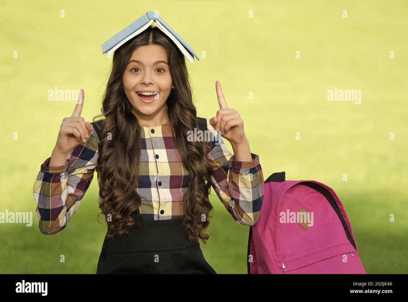 Joyeux petit enfant dans l'uniforme de l'école tenir le livre sur la tête pointant les doigts index vers le haut ensoleillé dehors, idée Banque D'Images