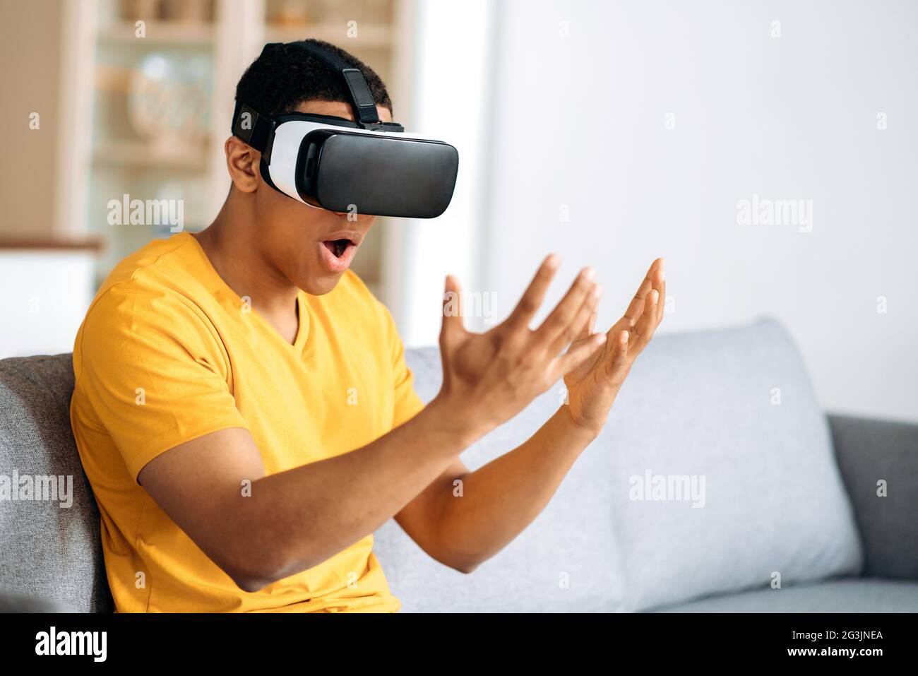Un homme hispanique moderne qui teste des lunettes VR. Un jeune homme utilise des lunettes de réalité virtuelle, regarde excité à ses mains, des gestes avec les mains, assis à la maison sur le canapé dans le salon Banque D'Images