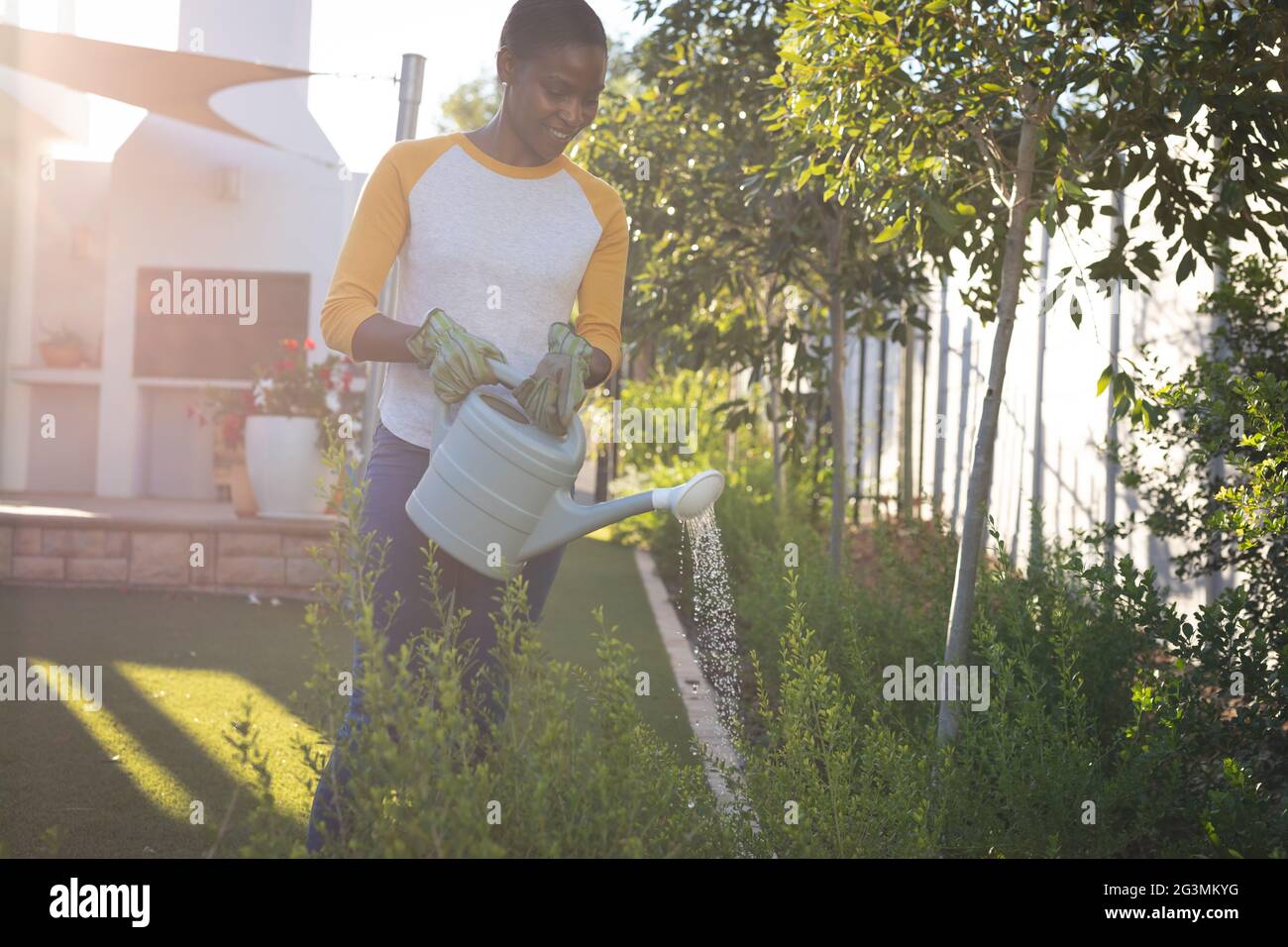 Souriante femme afro-américaine de jardinage, debout dans le jardin ensoleillé arrosoir plantes avec arrosoir Banque D'Images