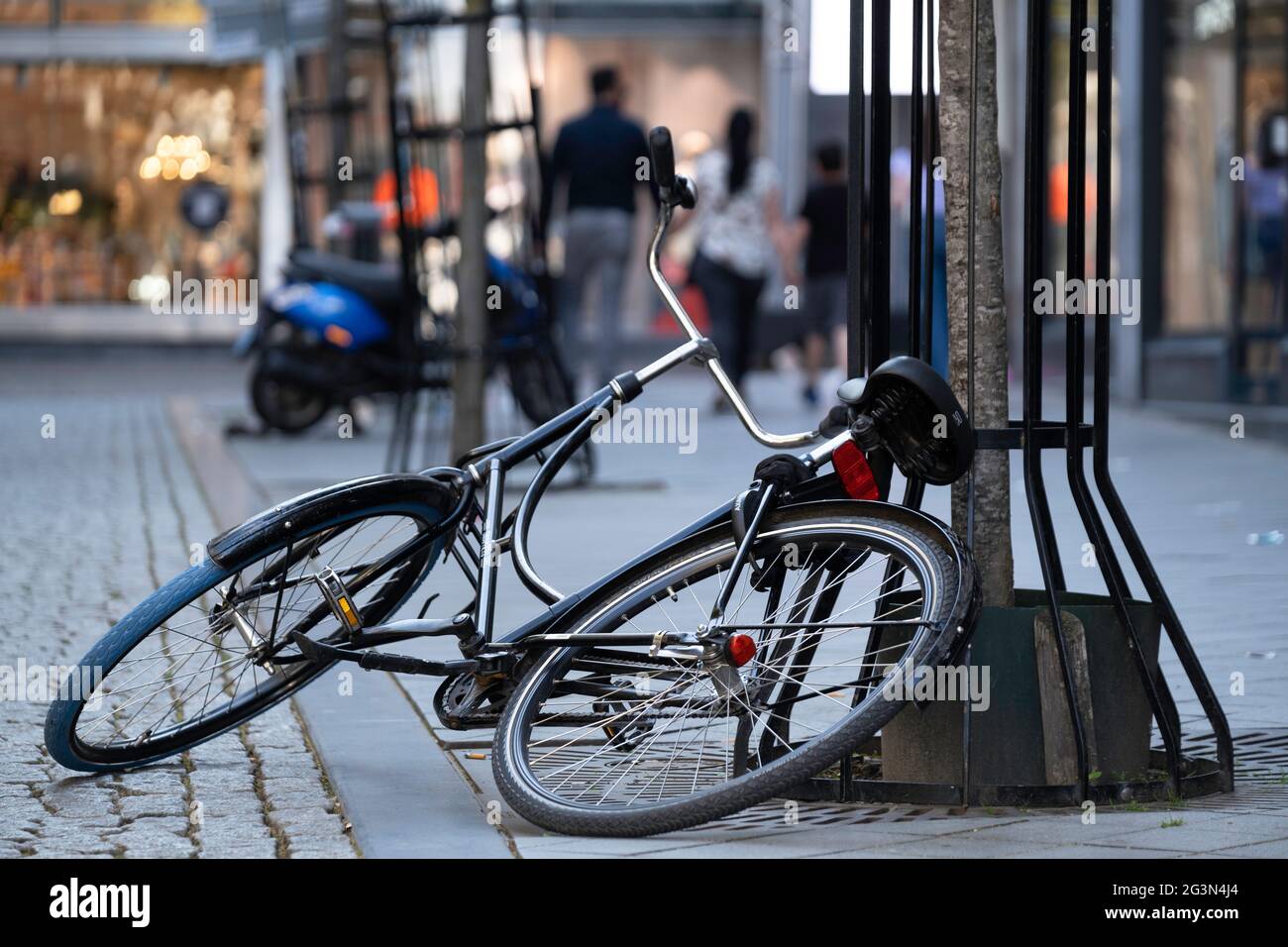 La bicyclette noire est à moitié sur le sol et à moitié contre un arbre dans une rue commerçante, garée sans prudence ou retournée. Vélo tombé dans la rue Banque D'Images