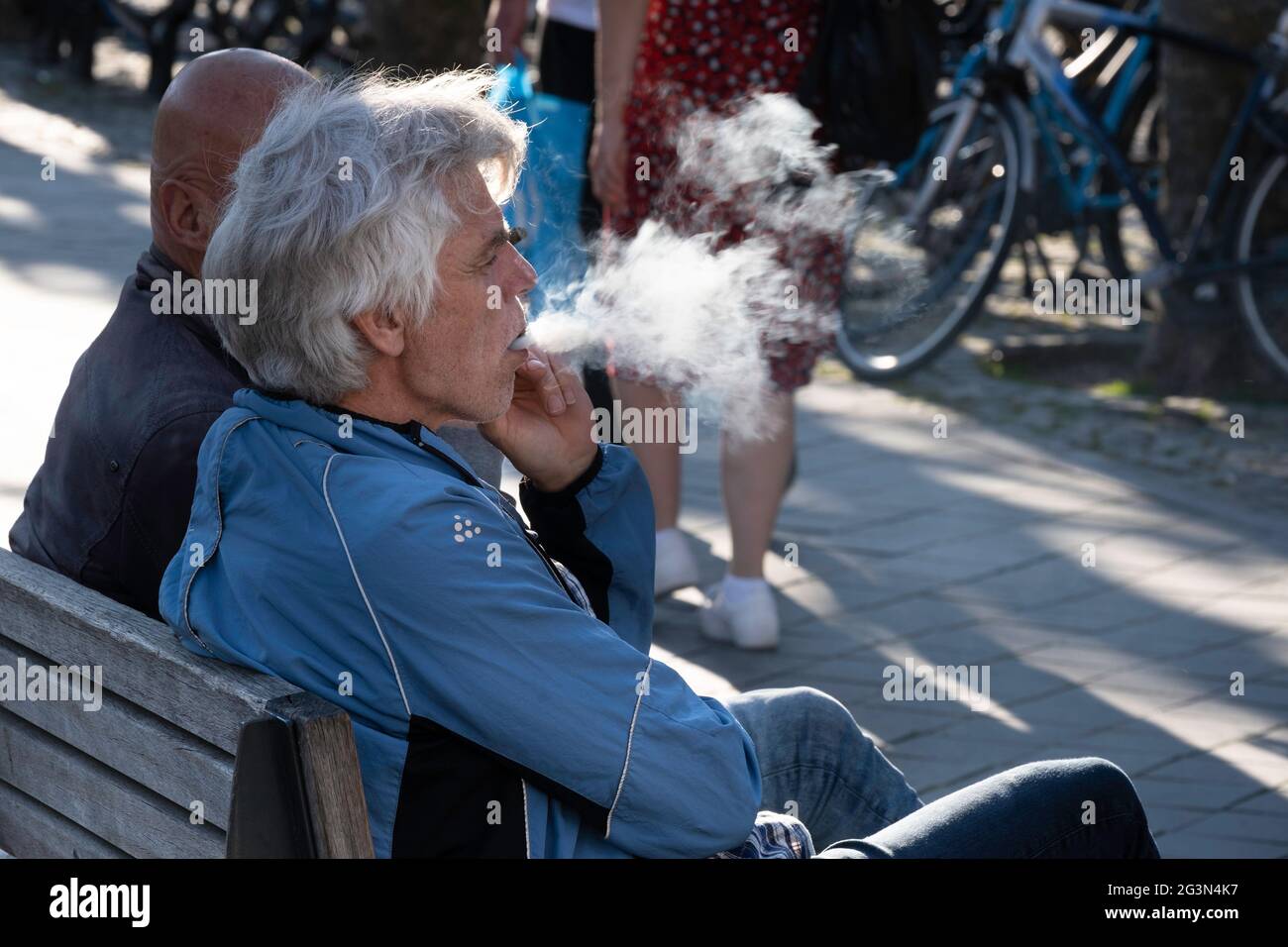 L'homme s'assoit sur un banc en bois, fume un cigare et souffle la fumée hors de sa bouche. Vue latérale avec la fumée comme un nuage blanc dans le rétroéclairage Banque D'Images
