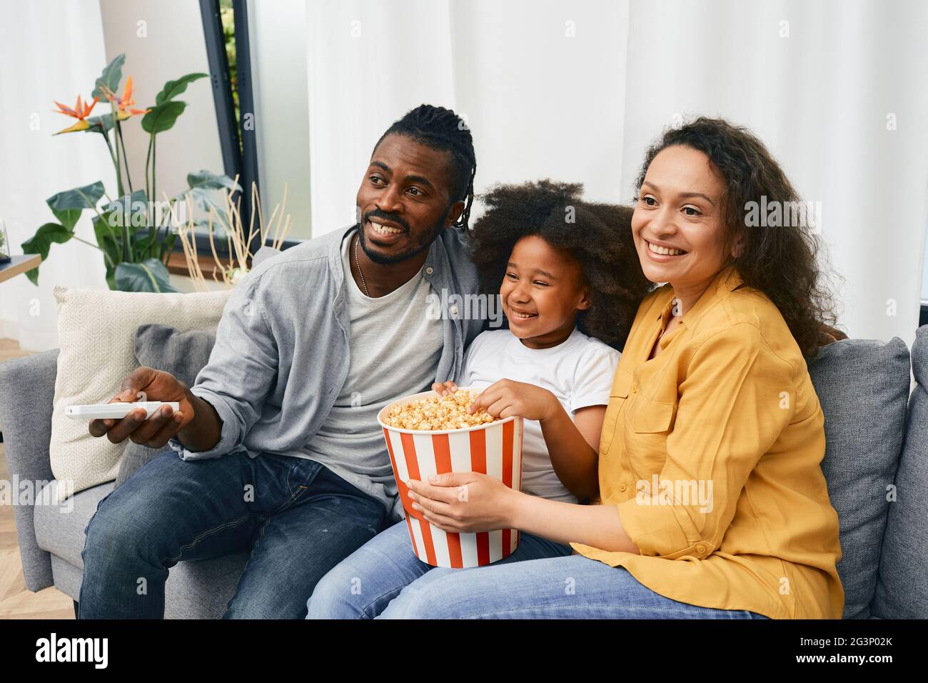 Une famille amusante avec une petite fille regarde un film de comédie ensemble et mange du pop-corn dans un grand seau à rayures dans une maison confortable Banque D'Images